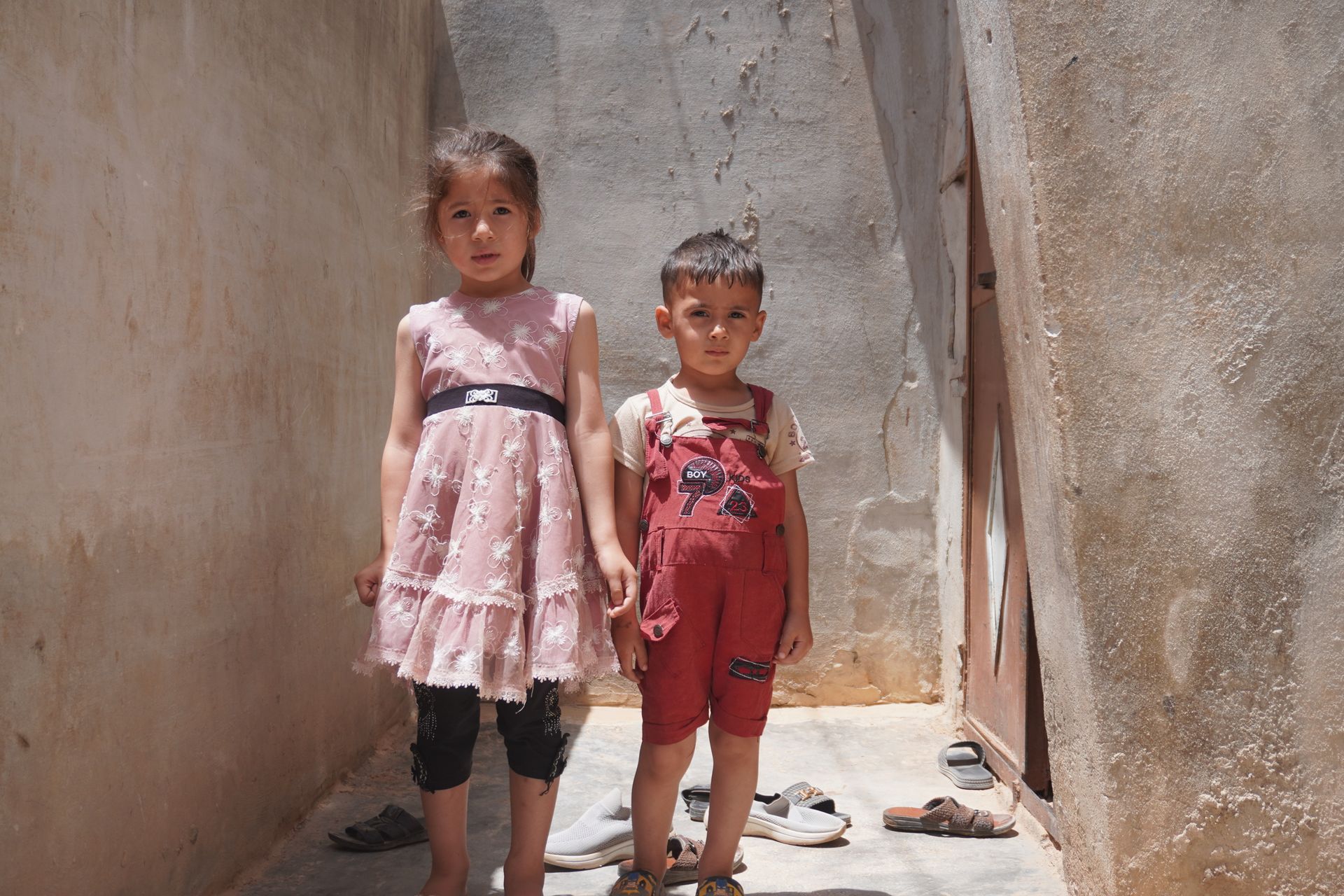 Deux enfants se tiennent côte à côte dans un couloir étroit en béton baigné de soleil.