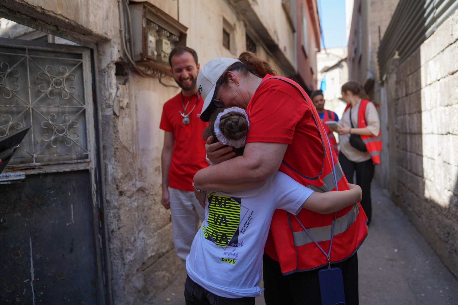 Une personne vêtue d'un gilet rouge enlace un enfant en chemise blanche. Deux autres personnes se tiennent dans un étroit passage extérieur à proximité.