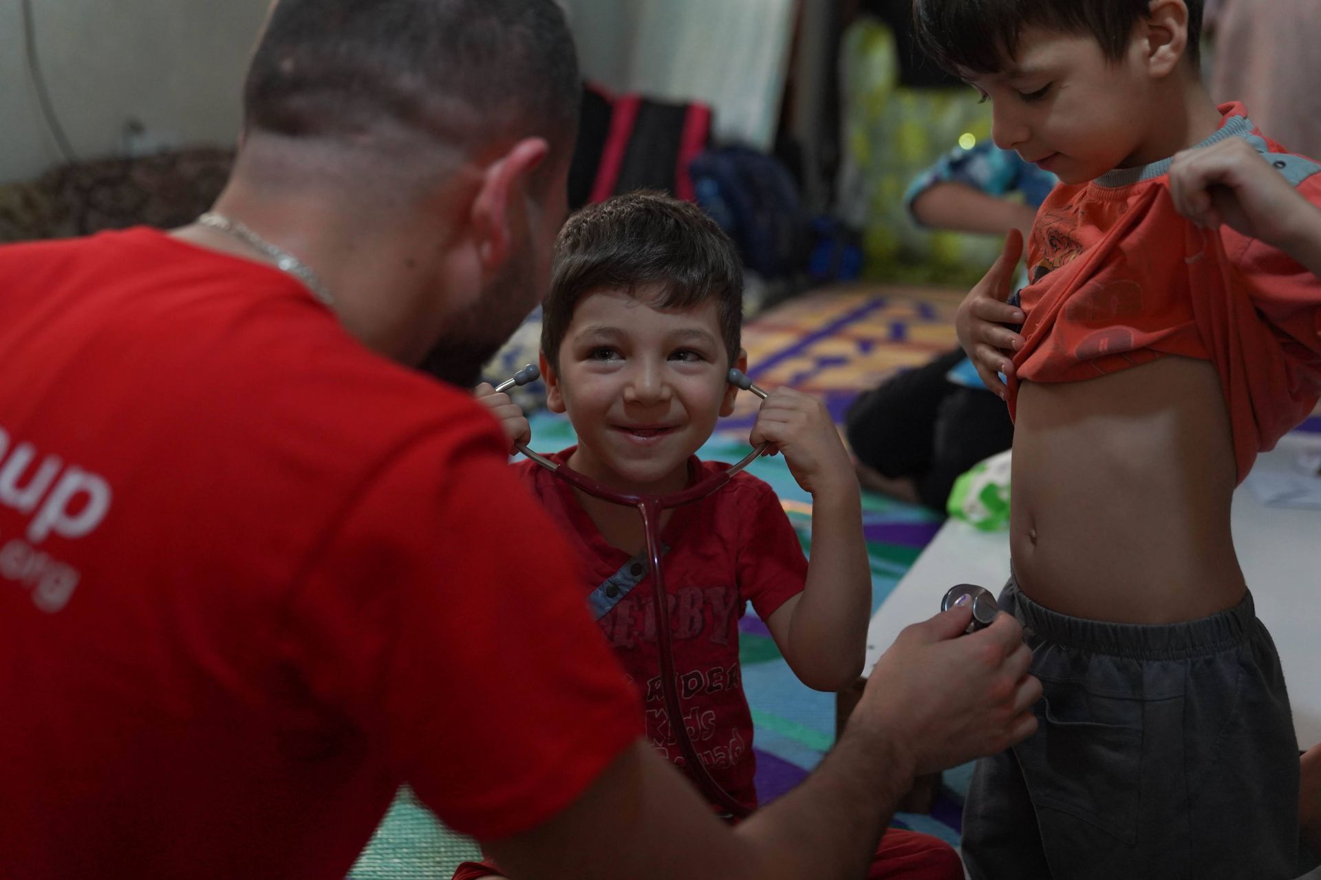 Dans un décor intérieur aux couleurs vives, un bénévole vêtu d'un t-shirt rouge examine l'abdomen d'un enfant souriant à l'aide d'un stéthoscope.