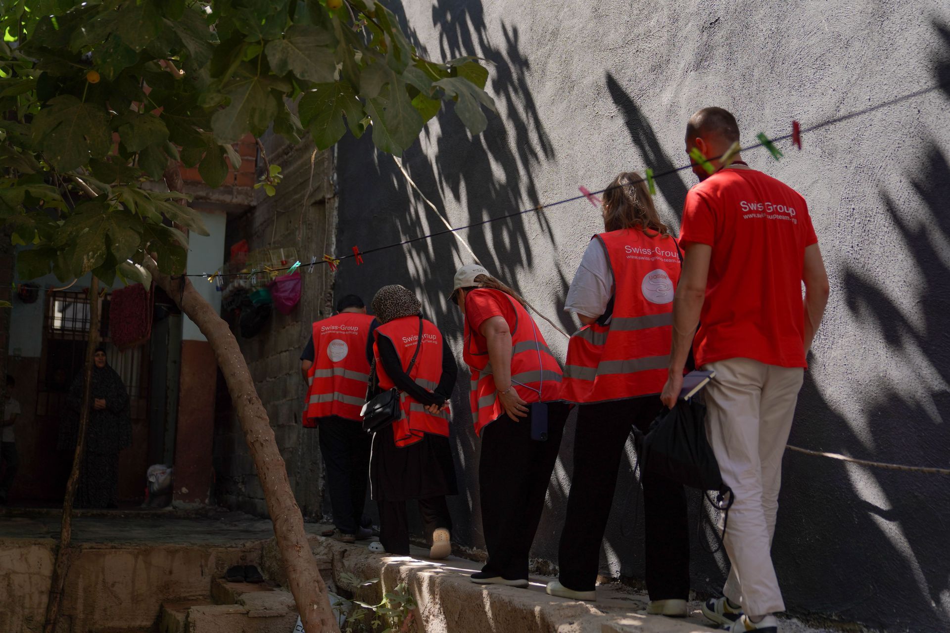 Quatre personnes vêtues de gilets et de chemises rouges de volontaires marchent en file indienne le long d'un mur extérieur, dans un passage étroit et ombragé.