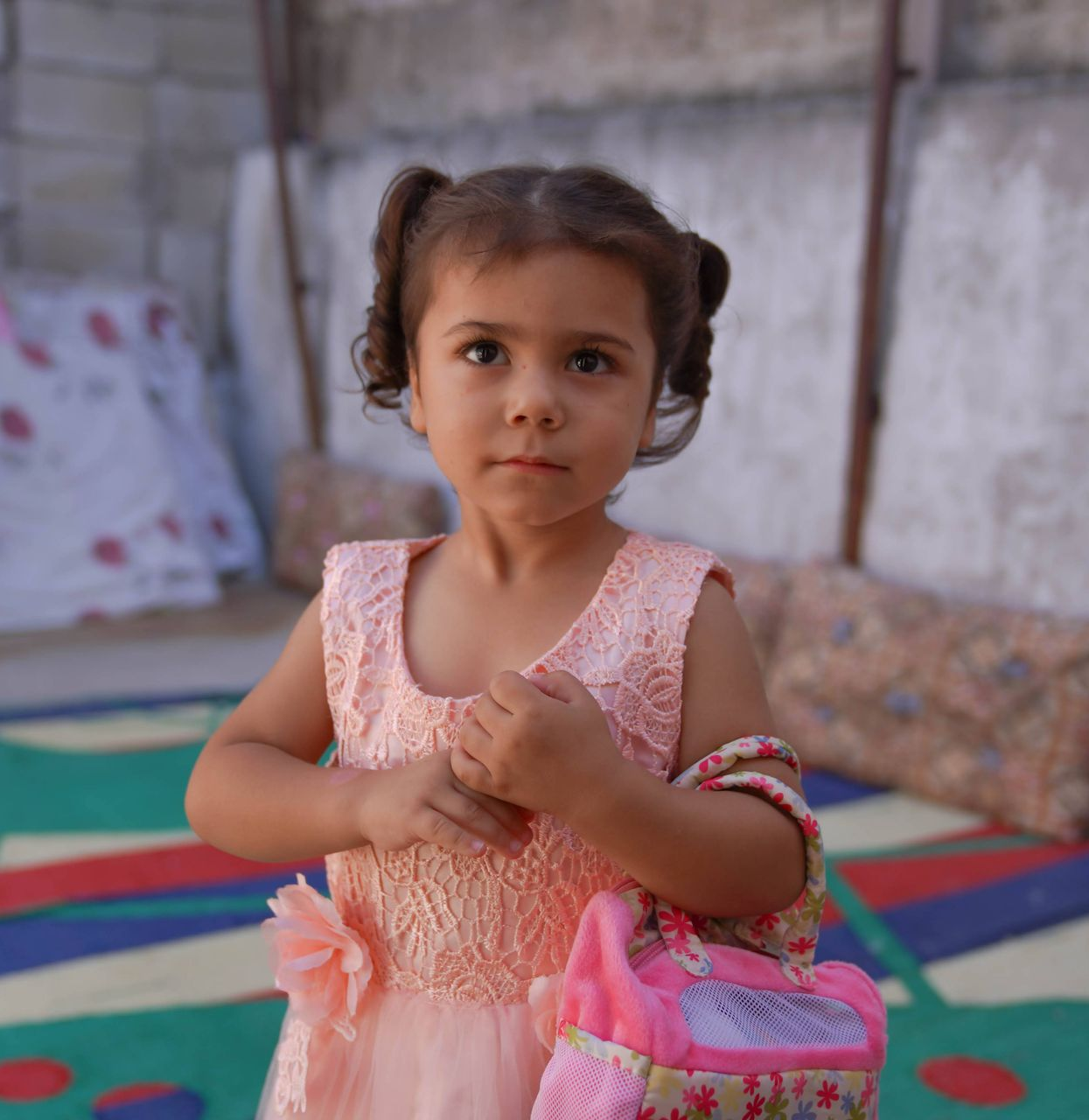 Une personne vêtue d'une robe rose pâle et coiffée de couettes jumelles tient un petit sac à fleurs, debout sur un tapis à motifs colorés.