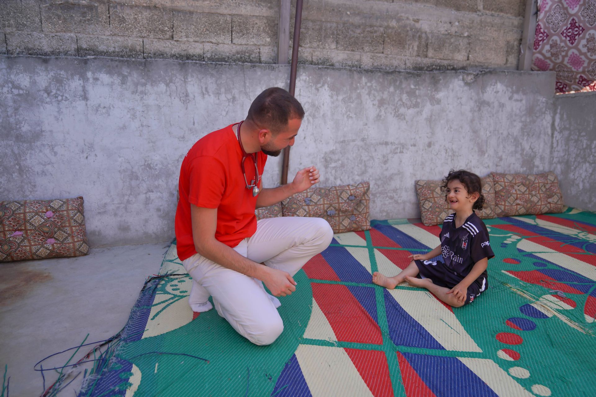 Une personne vêtue d'un t-shirt rouge est agenouillée sur un tapis à motifs pour interagir avec un enfant assis par terre près d'un mur en béton.