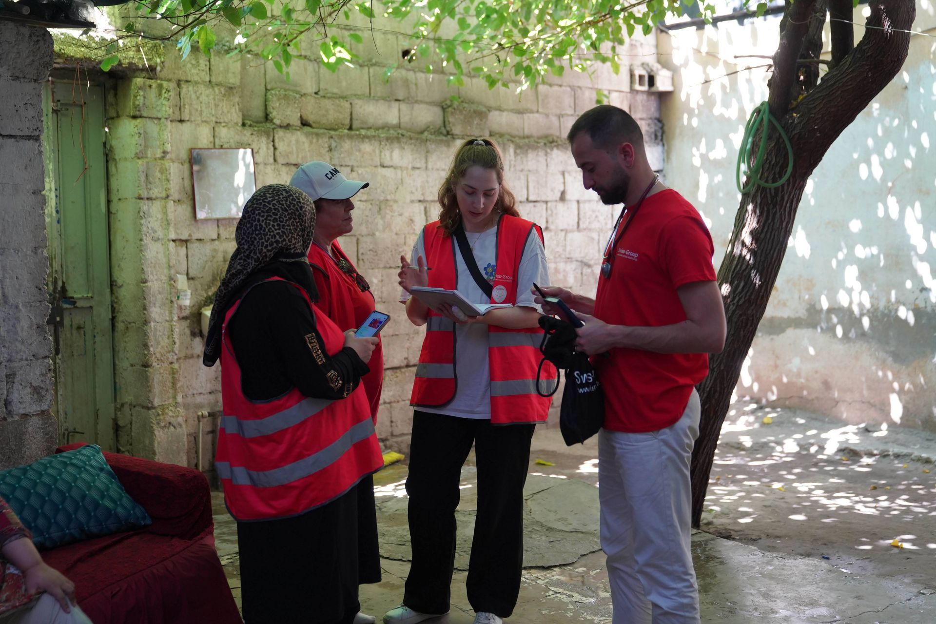 Quatre personnes vêtues de gilets rouges de volontaires consultent des notes, debout dans une cour en briques ombragée par des arbres.
