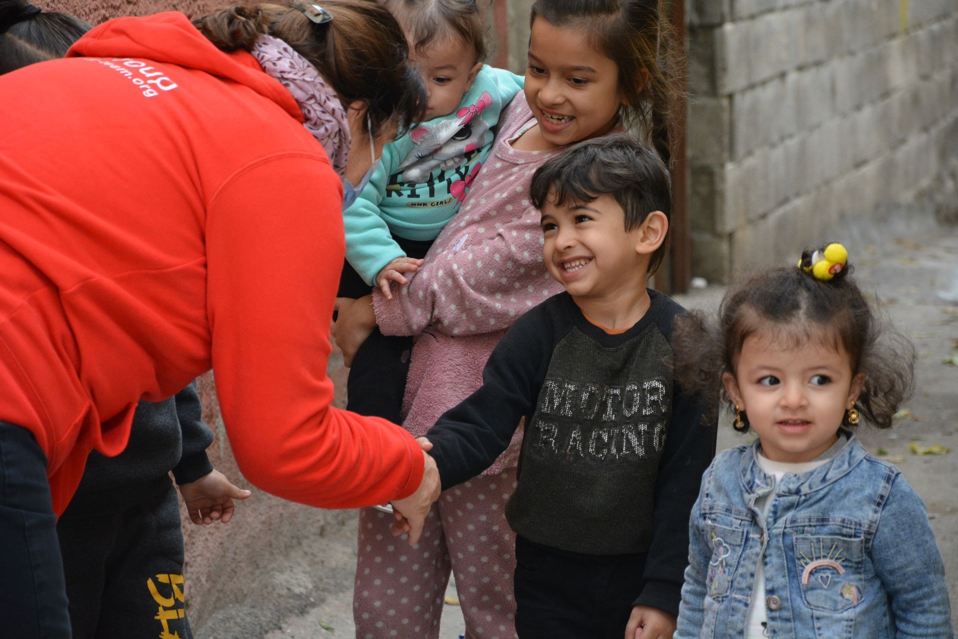 Une personne portant un sweat-shirt à capuche rouge serre la main d'un enfant souriant, entouré d'autres enfants en extérieur.