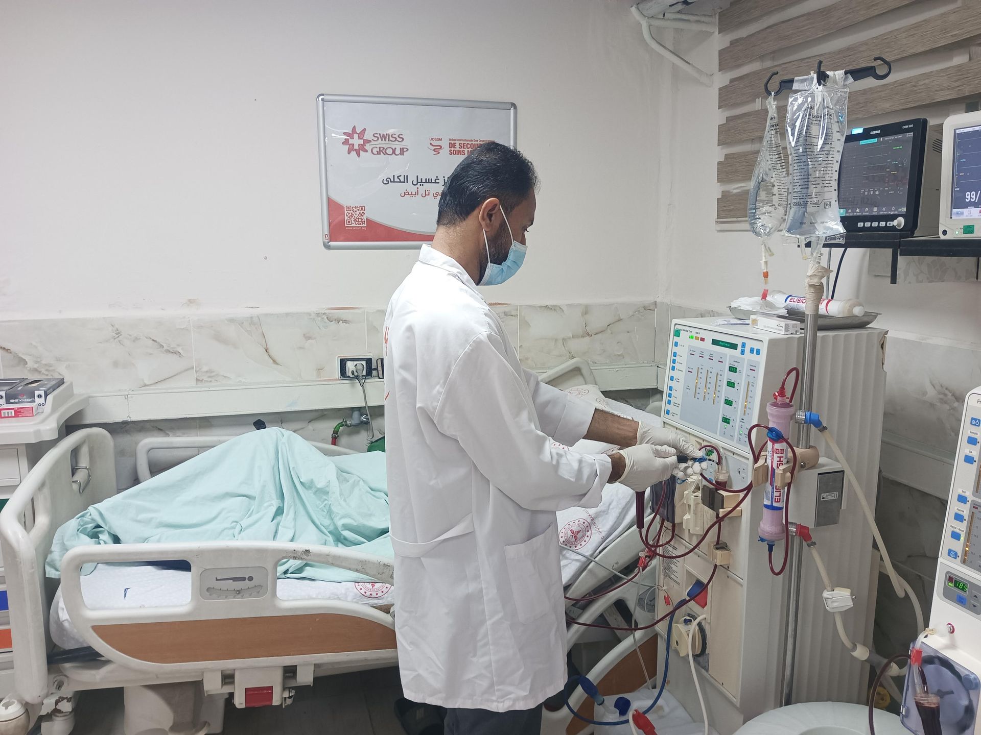 Un professionnel de santé en blouse blanche et masque actionne une machine de dialyse à côté d'un patient alité dans un hôpital.