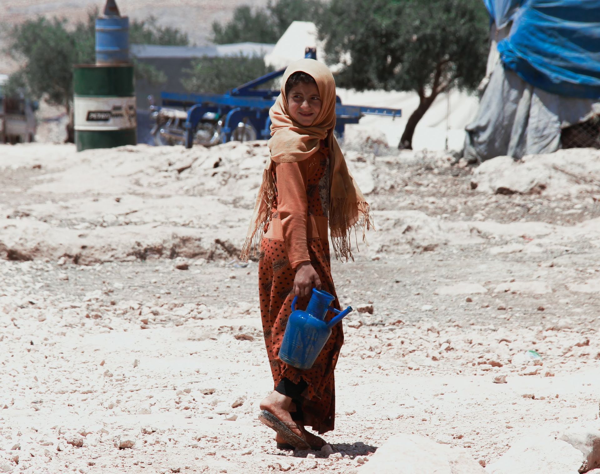 Une personne vêtue d'une robe à motifs et d'un foulard sur la tête traverse un paysage rocailleux en portant un récipient d'eau en plastique bleu vif.