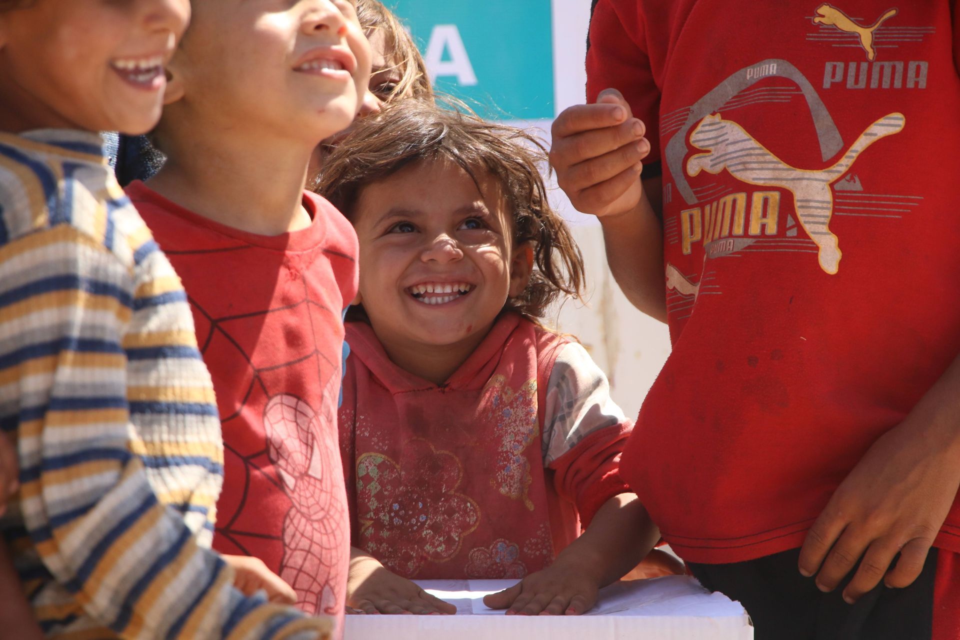 Un enfant souriant se tient au milieu d'autres personnes en vêtements décontractés, avec un panneau flou visible en arrière-plan.