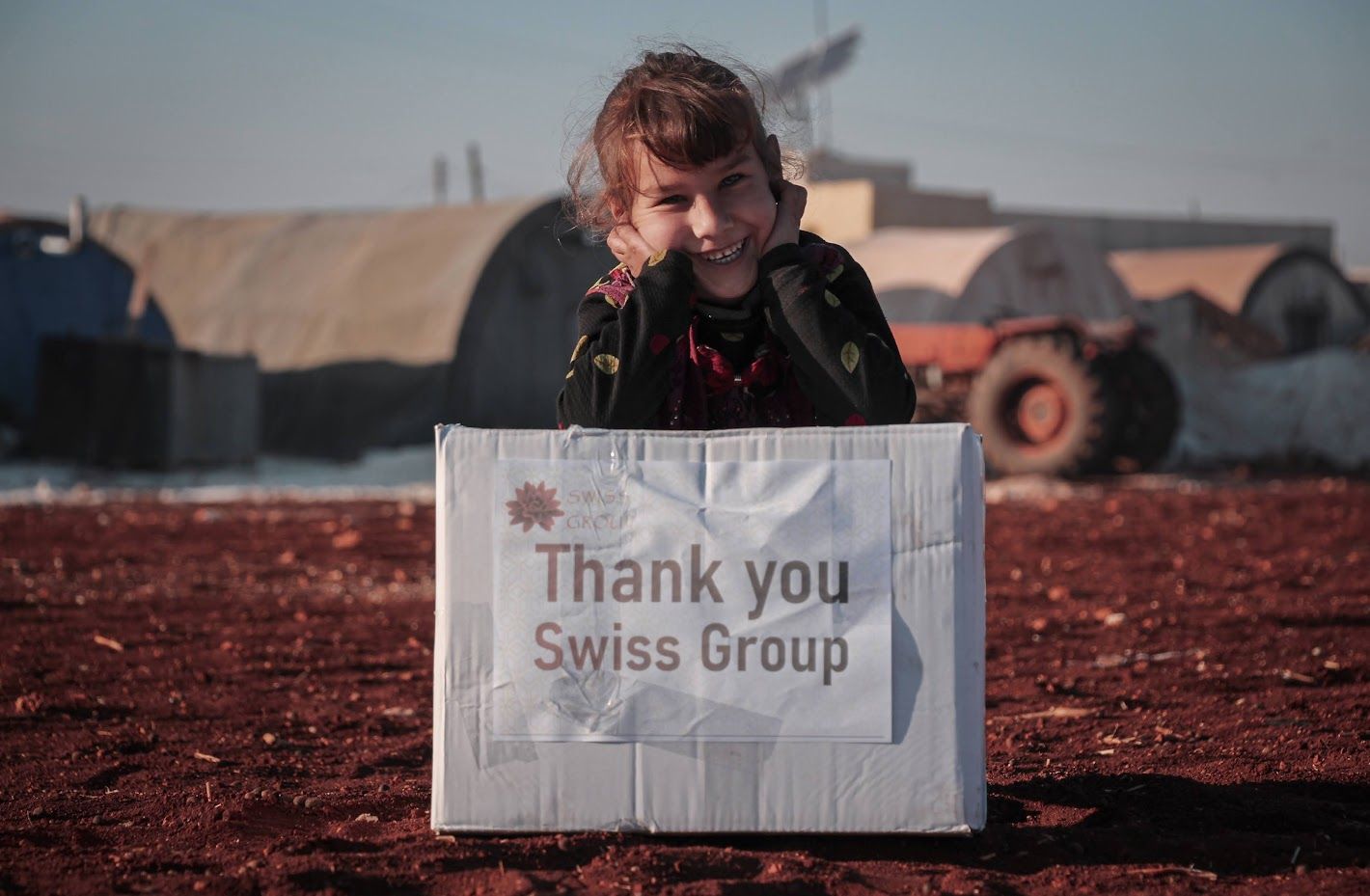Une personne souriante, appuyée sur un carton où l'on peut lire « Merci Swiss Group », se dresse devant de grandes tentes industrielles en forme d'arche.