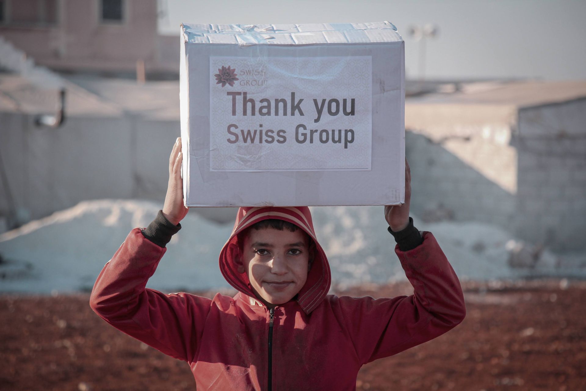 Dans un espace extérieur poussiéreux, une personne portant une veste à capuche rouge tient au-dessus de sa tête une boîte étiquetée « Thank Swiss Group ».