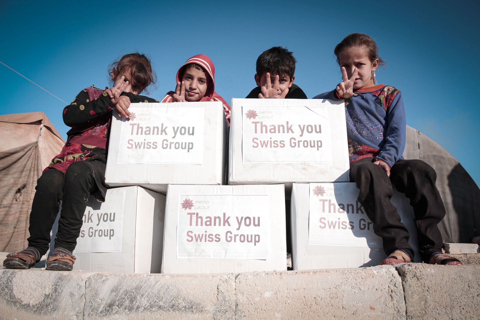 Quatre enfants sont assis dehors, derrière des boîtes étiquetées « Merci Groupe Suisse », faisant des signes de paix face à la caméra.