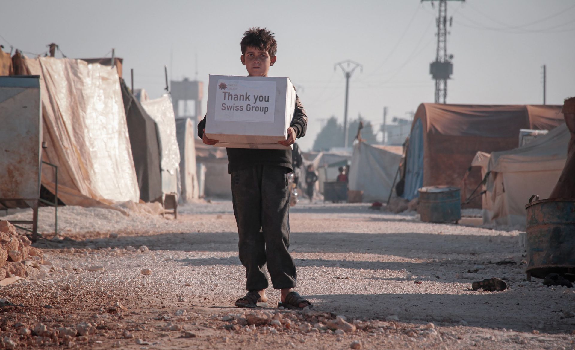 Une personne se tient debout dans un camp de réfugiés, tenant une boîte étiquetée « Merci Turquie-Syrie ».
