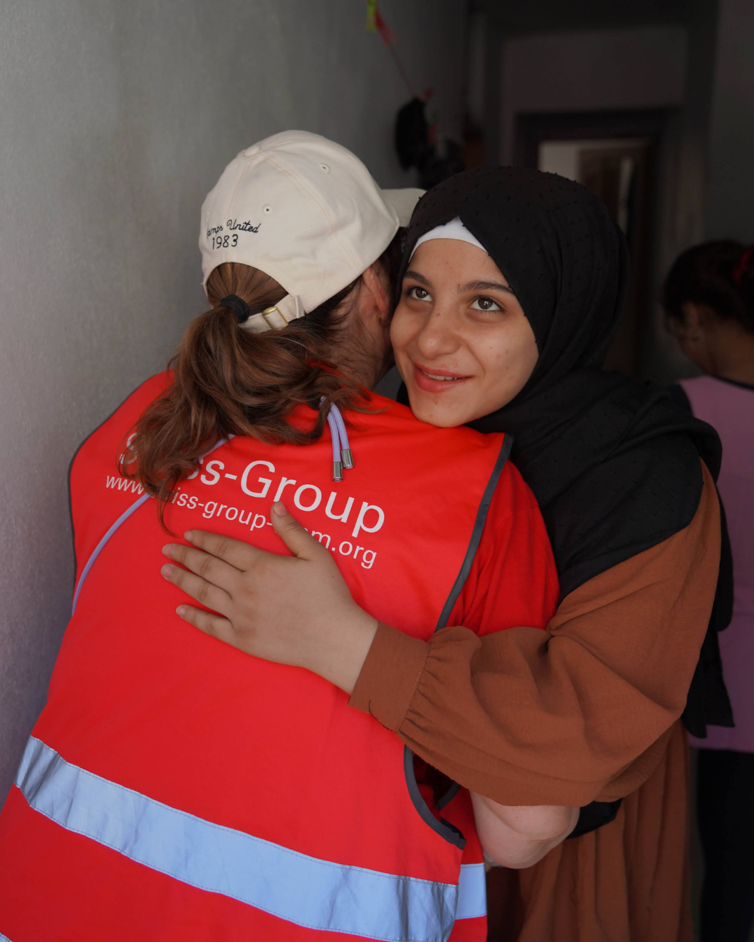 Une personne portant un gilet rouge avec la marque « is-Group » enlace une autre personne portant un foulard noir et une chemise beige.