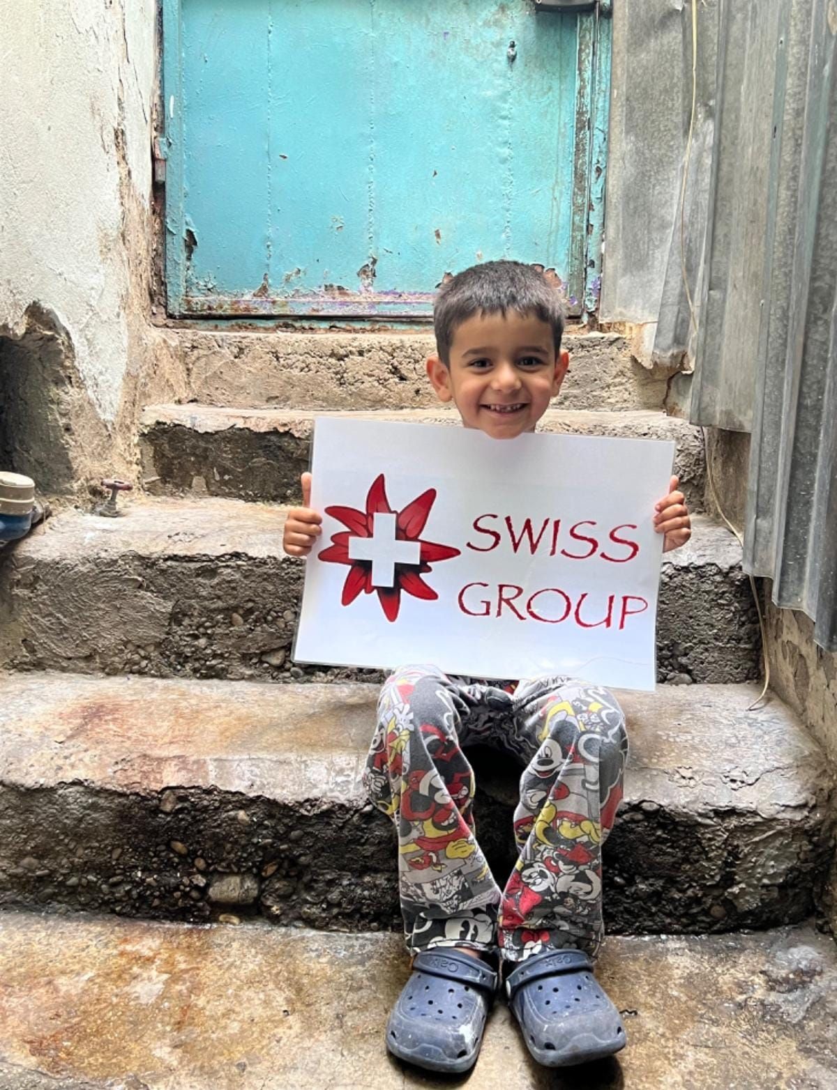 Un enfant souriant est assis sur des marches en pierre à l'extérieur, tenant une pancarte blanche sur laquelle on peut lire « SWISS GROUP » avec un emblème rouge et blanc.