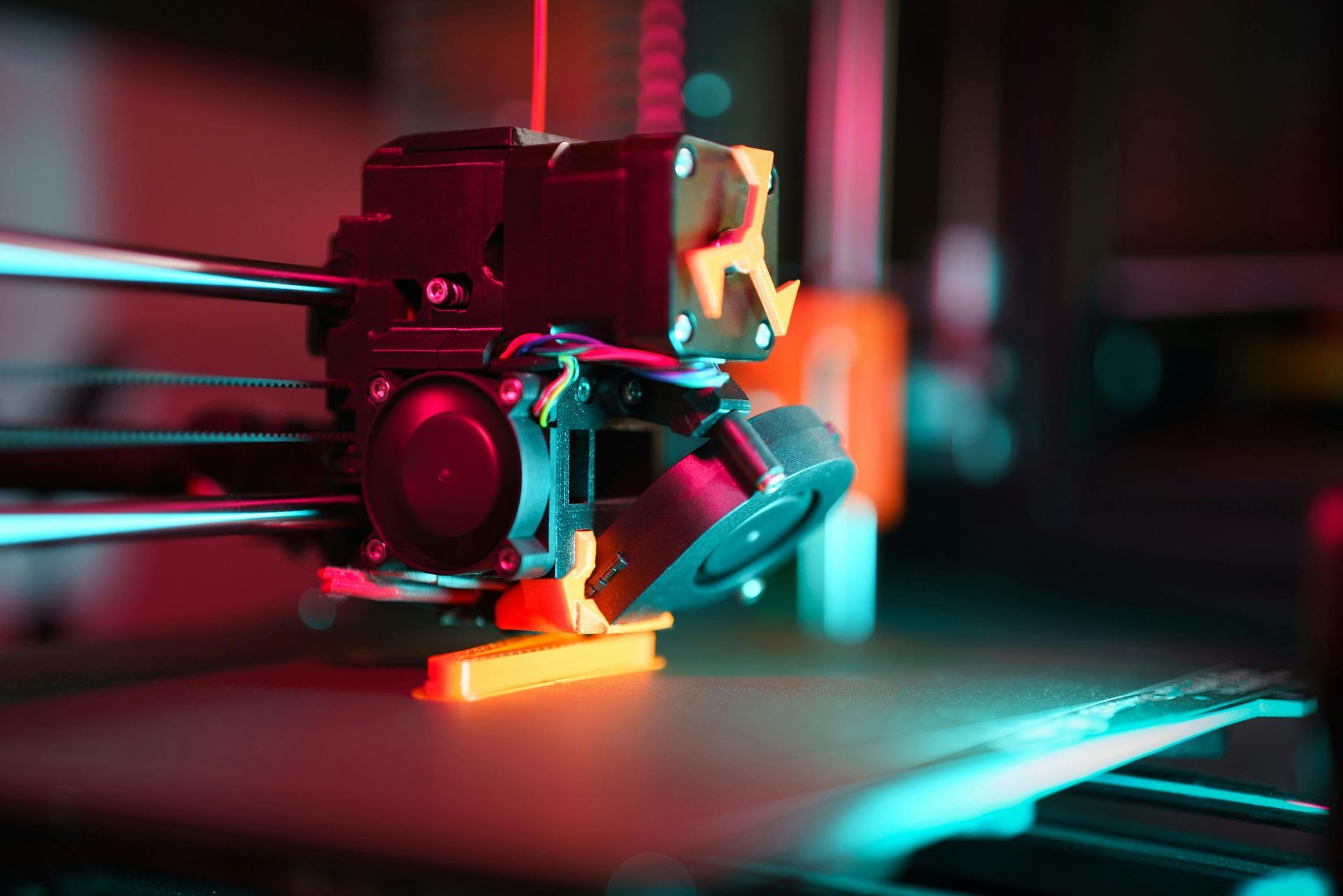 3D printer nozzle extruding orange plastic onto a bed, with red and blue lighting.