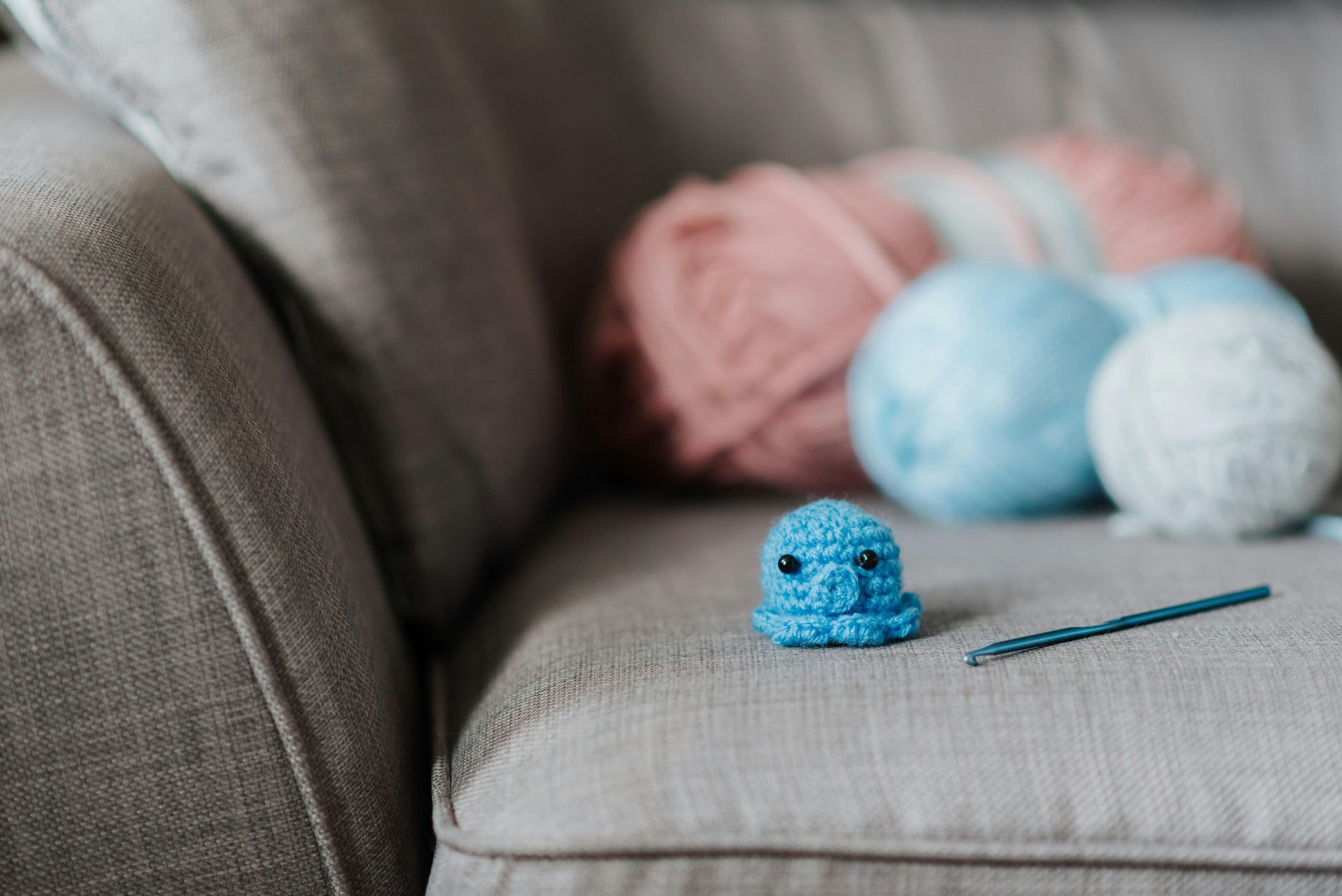 Small blue crocheted octopus and hook on a gray couch; yarn balls in pink, light blue, and white in background.