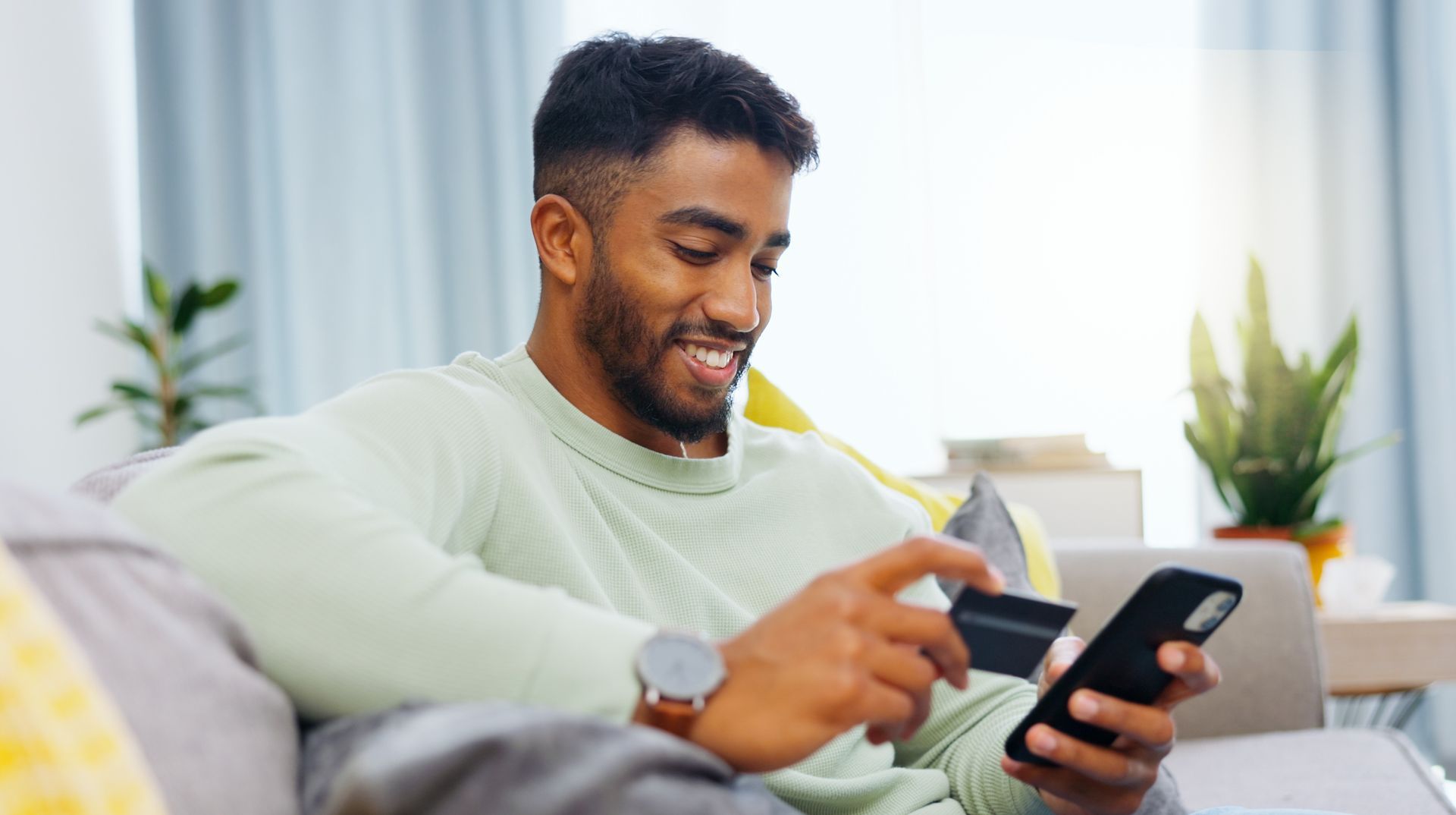 a man is smiling and sitting on a sofa using a credit card and a mobile phone