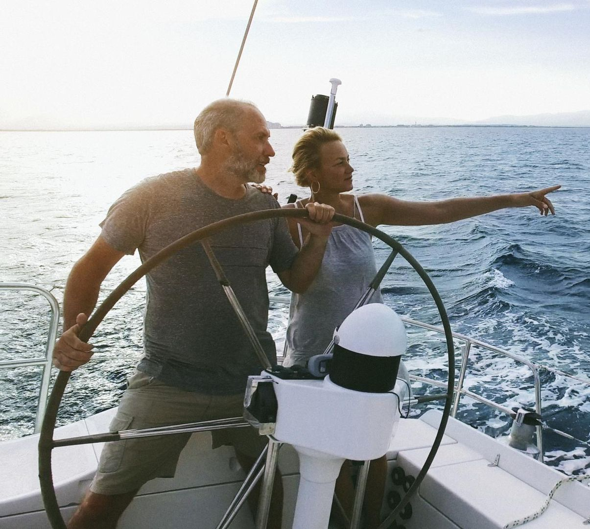 Man steers sailboat, woman points towards ocean horizon.