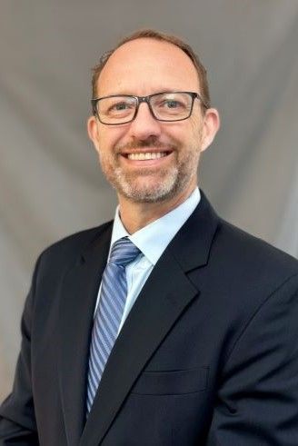 Man in glasses and suit, smiling at the camera against a neutral gray background.
