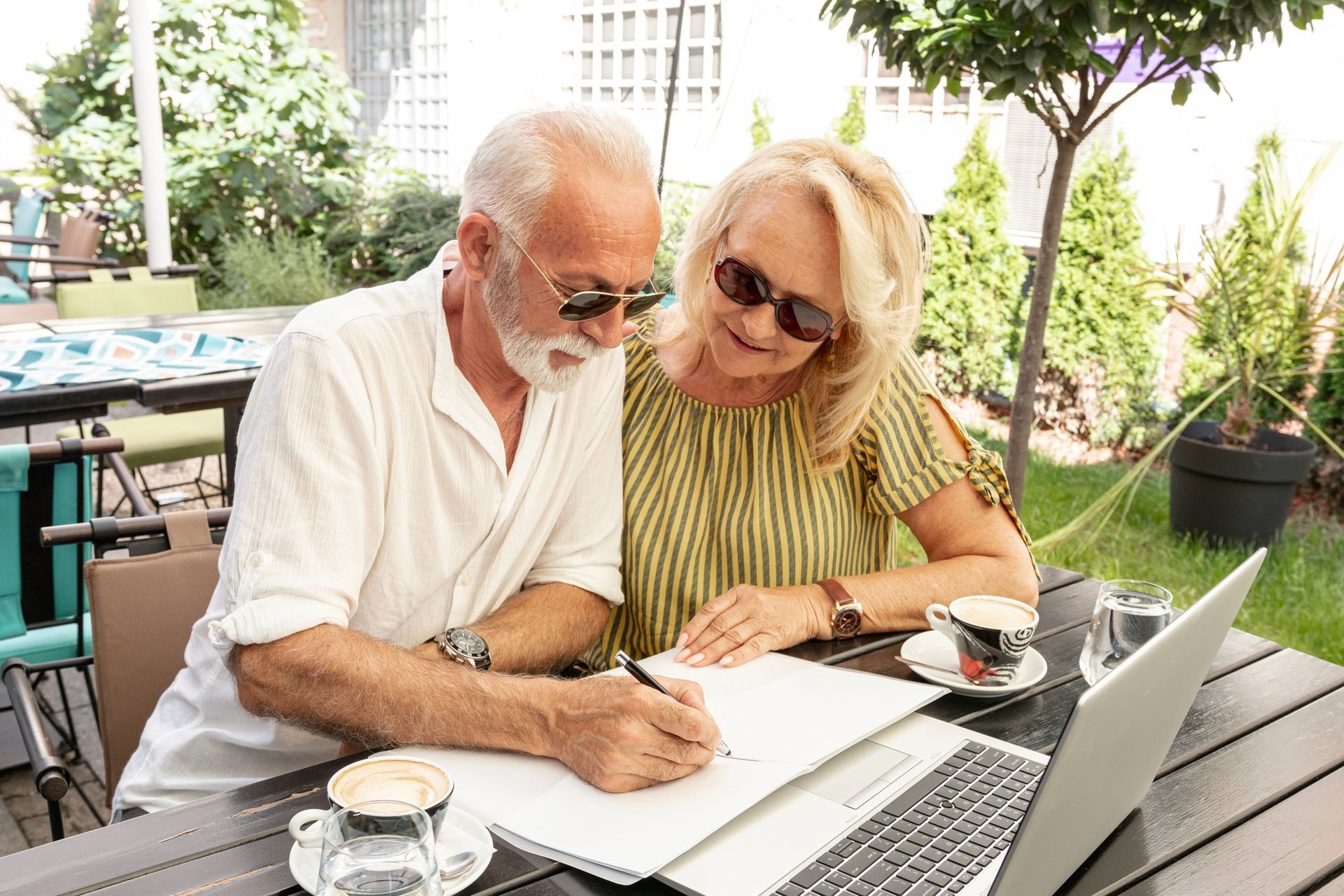 Couple at table, man writing on paper, woman watches, coffee, laptop, outdoor cafe.