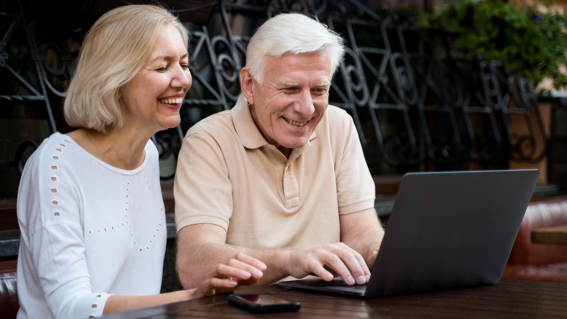 Couple smiles while looking at a laptop computer. Outdoors, sunny day.
