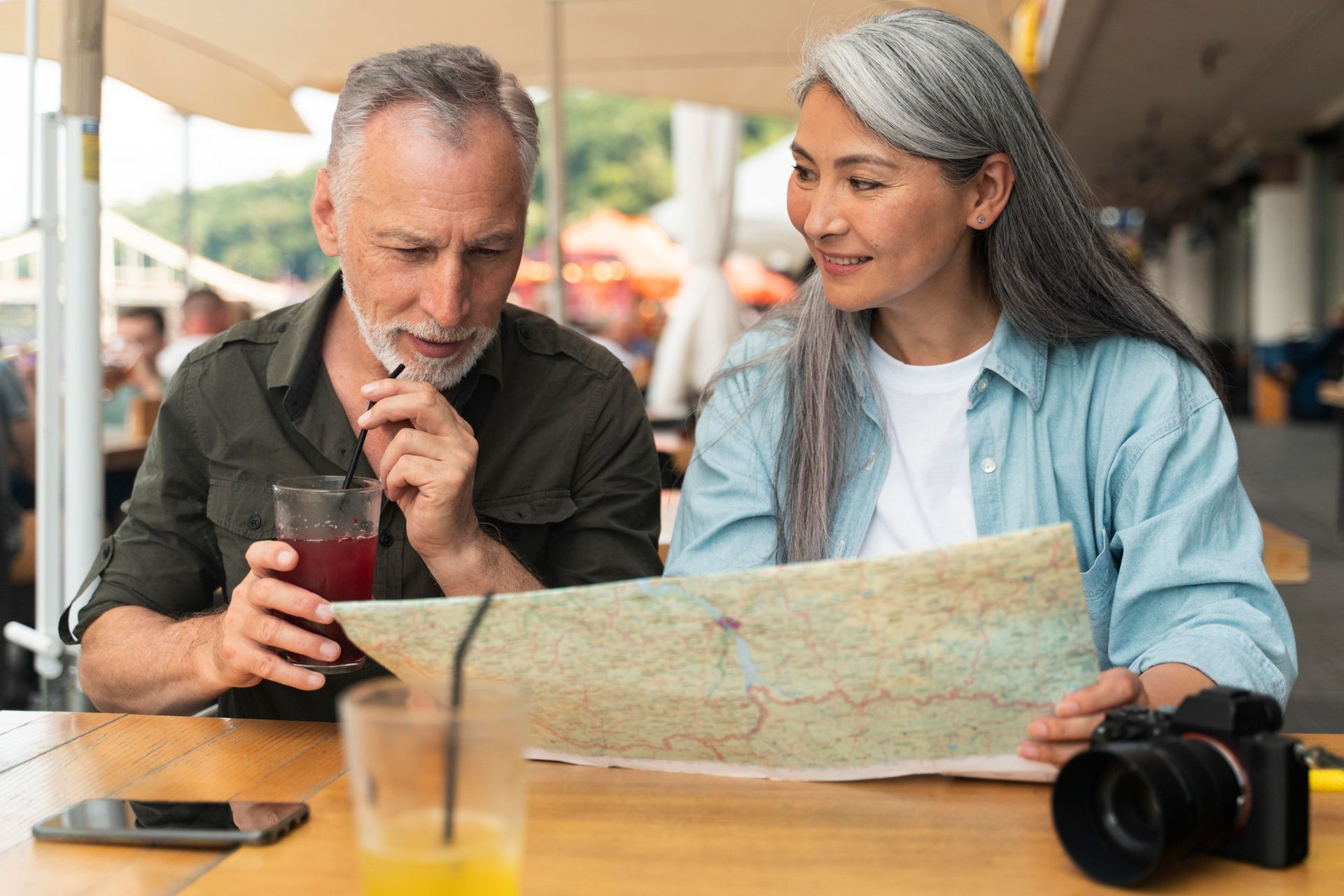 Couple at outdoor table looking at map, man drinking from a glass, camera.