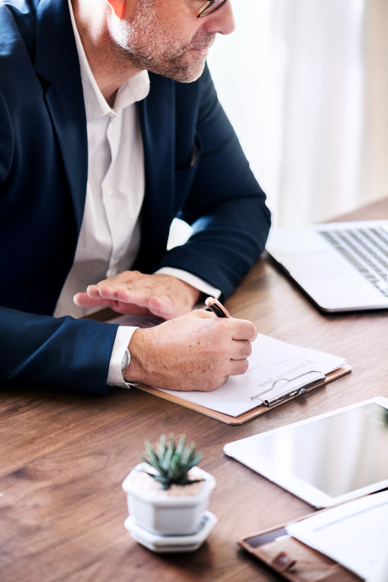 Man in suit writing on clipboard at a wooden table with laptop, tablet, and small plant.