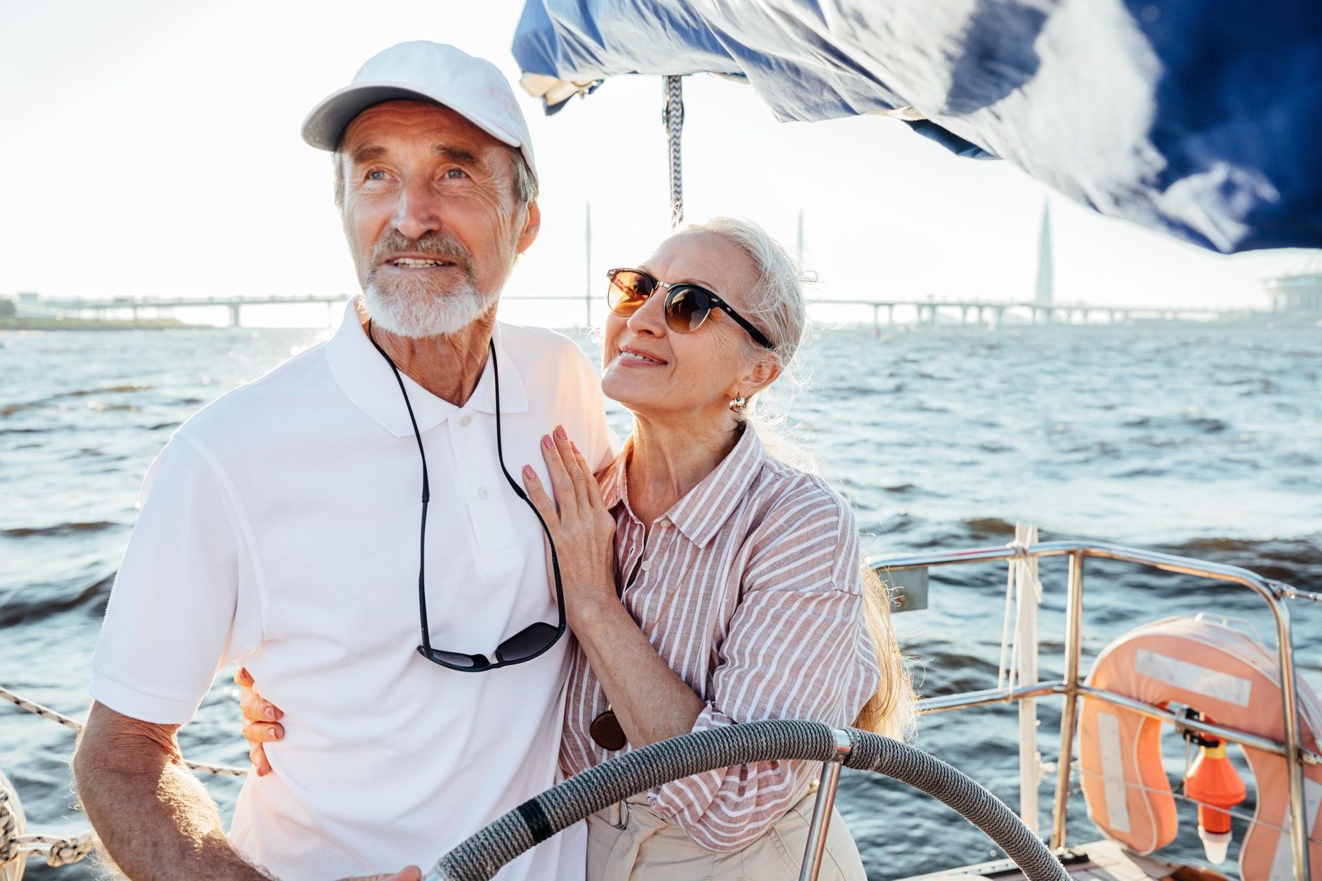 Couple on a sailboat, man at helm, woman with arm around him, ocean view, sunny.