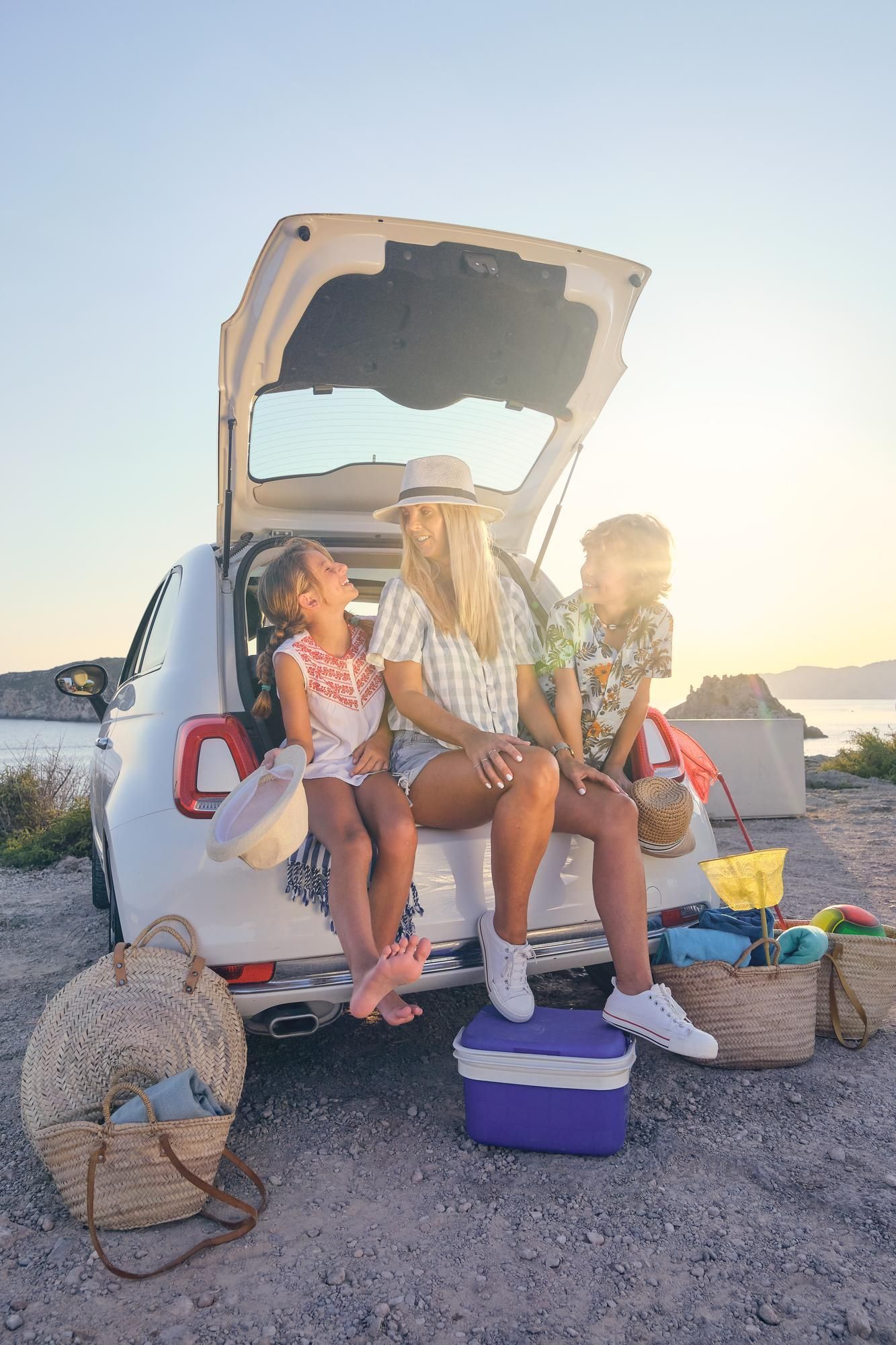 Woman and children sitting in car trunk, beach picnic setup, sunny day.