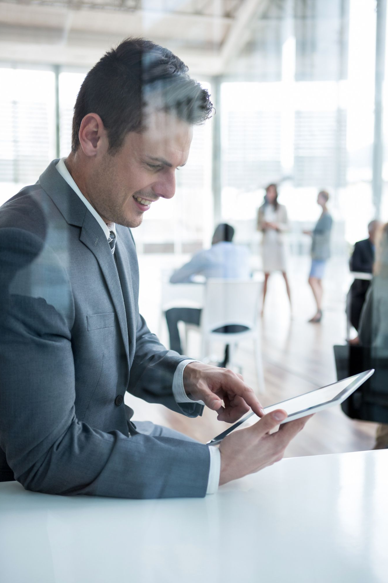Man in suit using tablet, smiling, in modern office setting with people blurred in background.