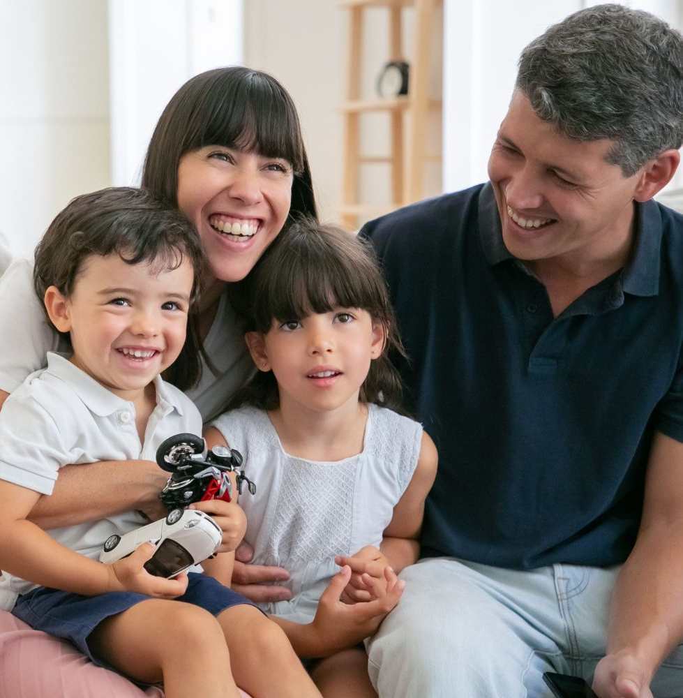 Family of four smiling, sitting on a couch. Children holding toy, mother and father looking at them.