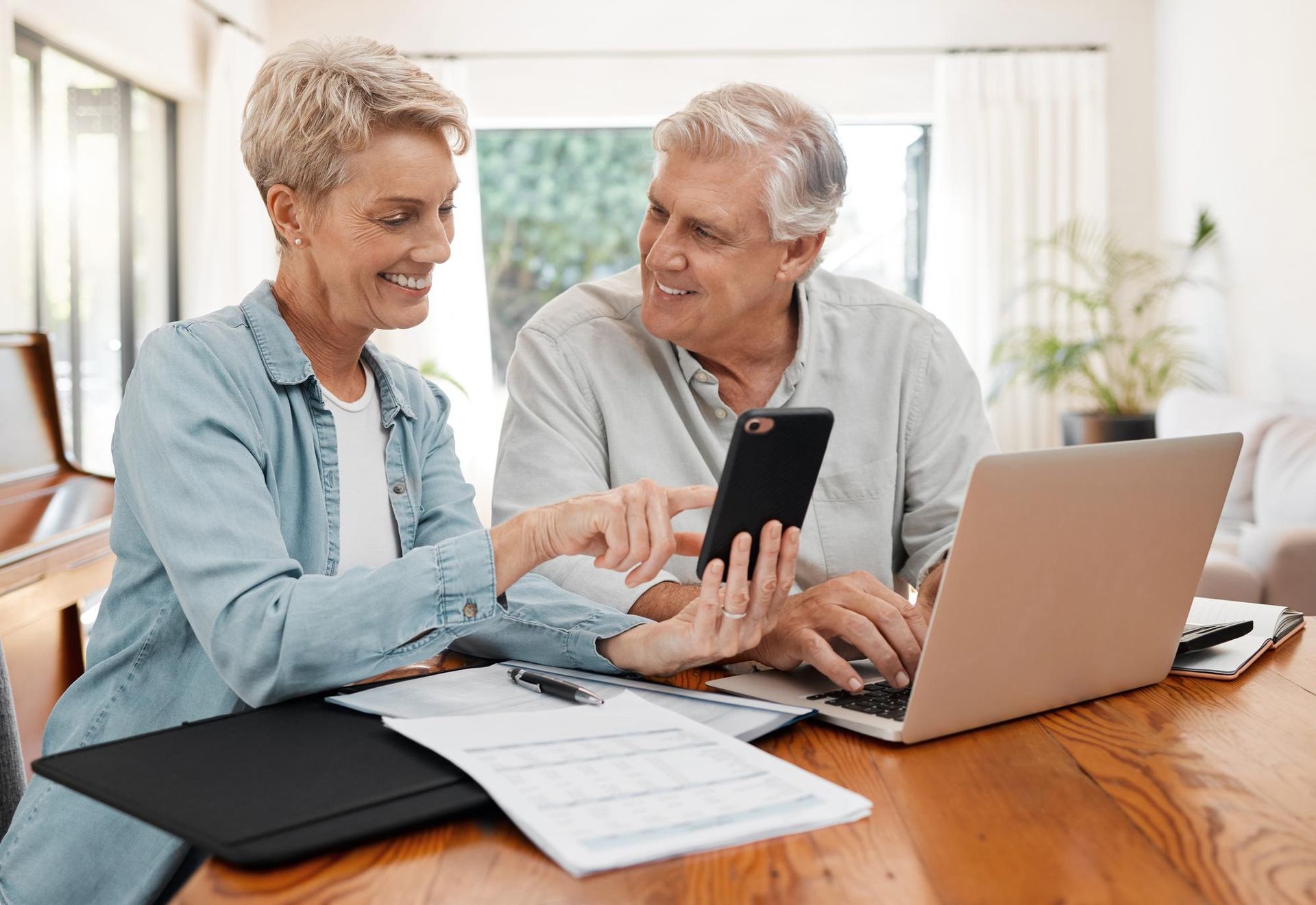 Older couple looking at phone together, laptop and documents on table.