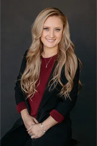 Woman with long blonde hair, smiling, wearing a burgundy shirt and black blazer, sitting against a gray background.