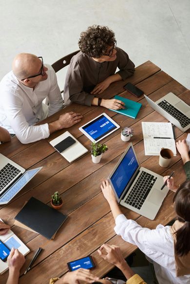 People at a wooden table in a meeting, with laptops, tablets, and notebooks; discussing work.