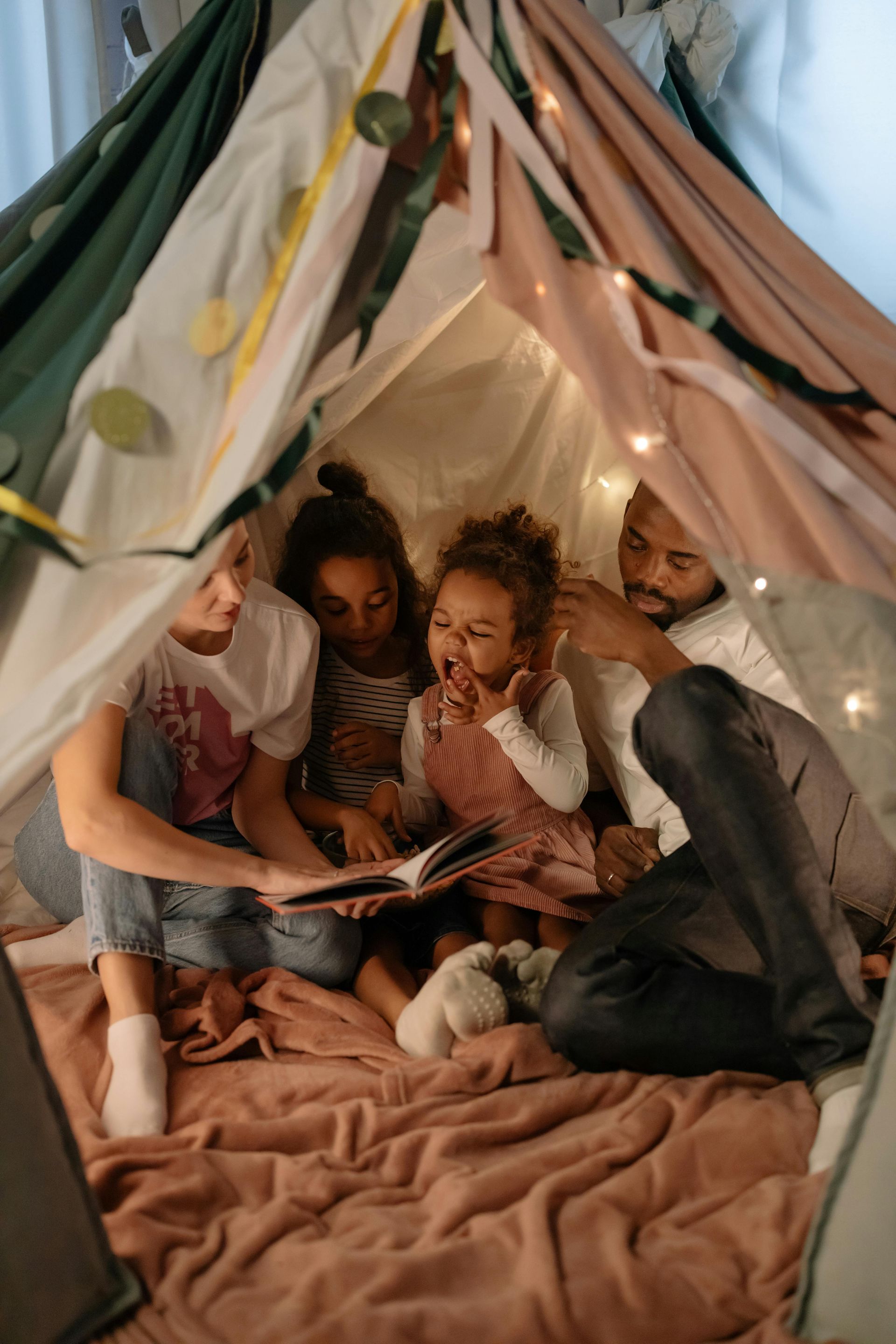 Family reading a book together inside a colorful blanket fort, lit with fairy lights.