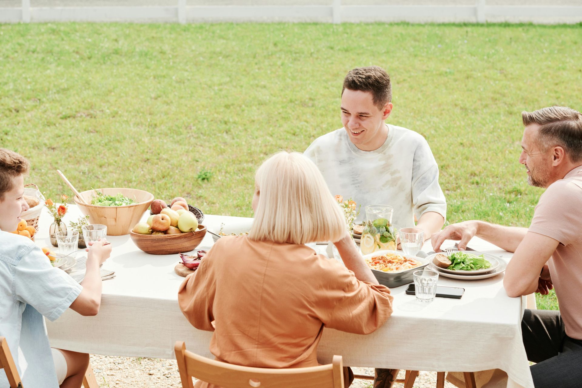 Family seated around a table, eating and talking outdoors on a sunny day.