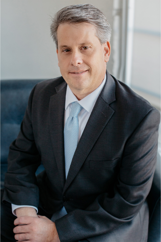 Man in dark suit, light blue tie, seated indoors, smiling at the camera.