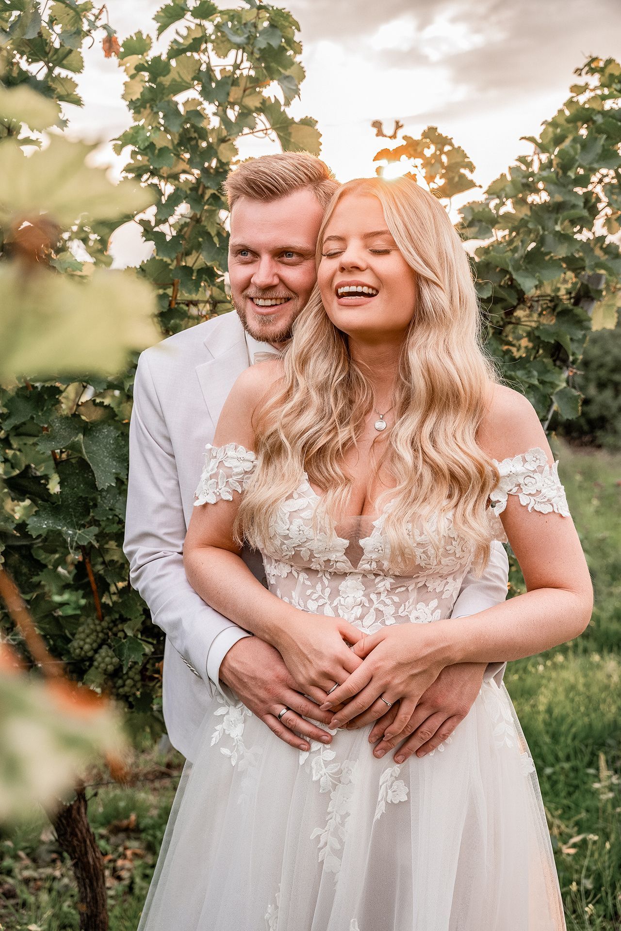 Bräutigam umarmt Braut, schaut in die Ferne, sie lacht mit geschlossenen Augen – Weinberge um sie herum