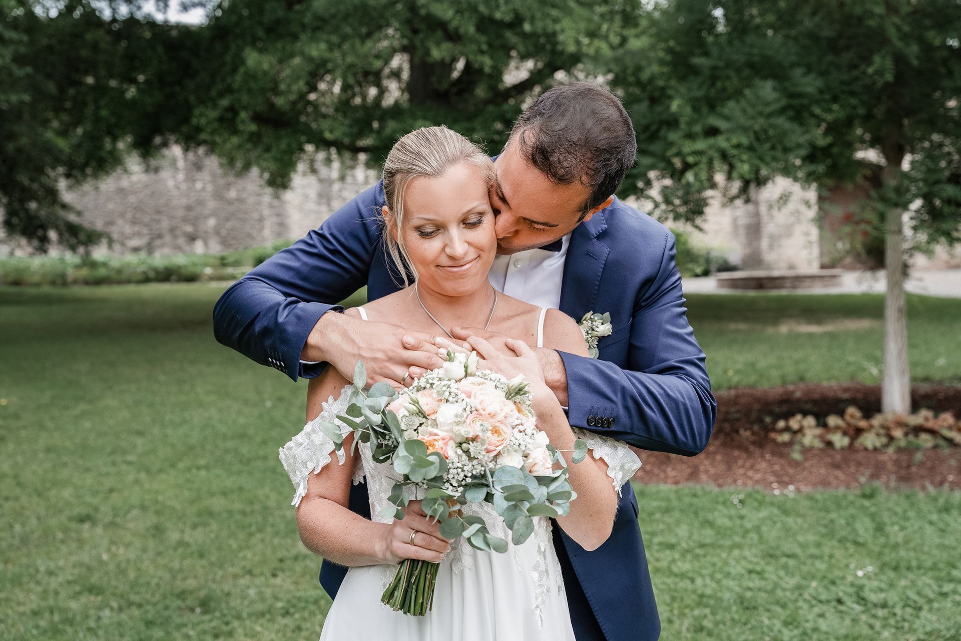 Bräutigam küsst die Braut auf die Wange, sie hält Brautstrauß in warmen Farben und schaut glücklich nach unten