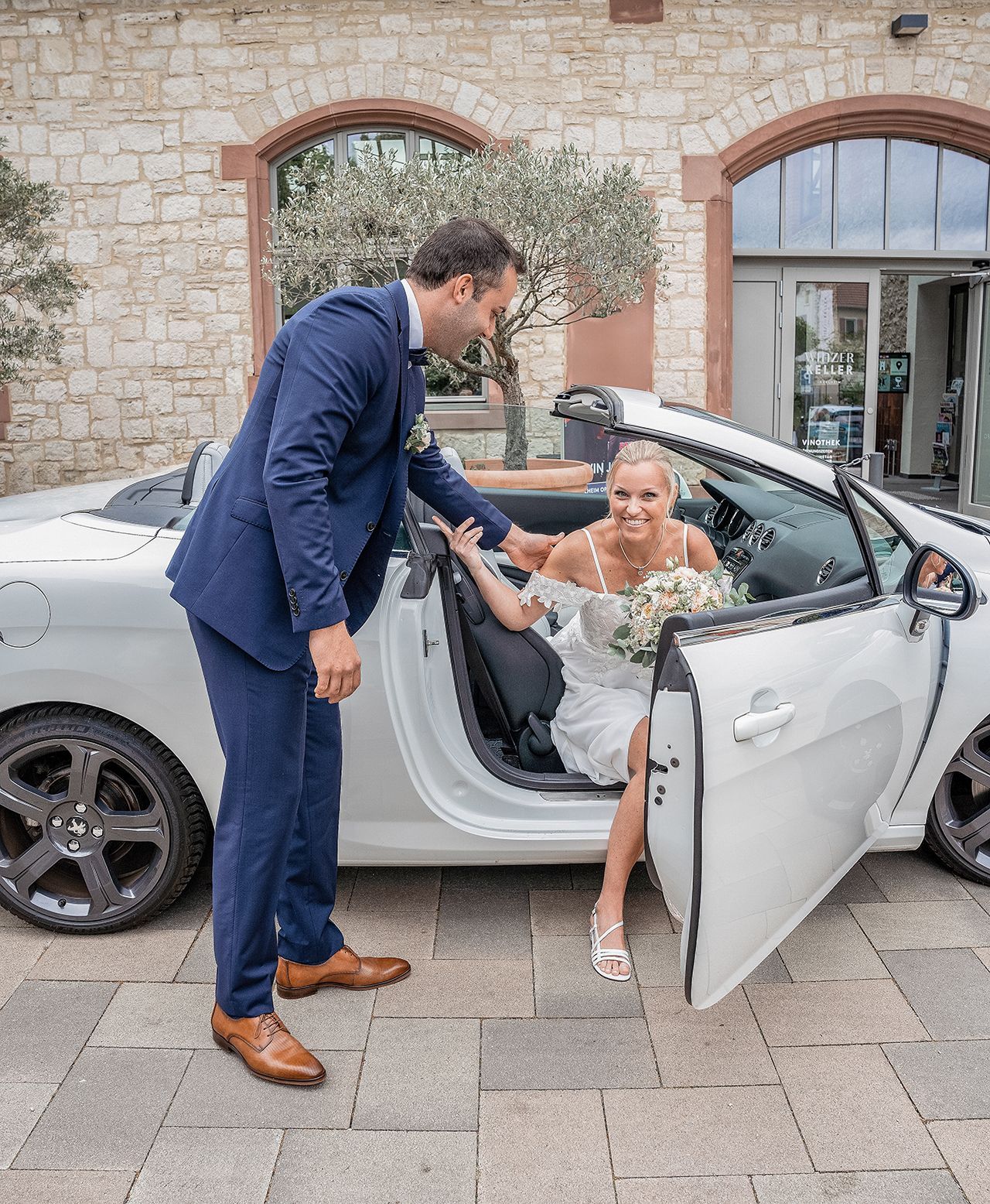 Braut steigt aus dem Brautauto, schaut in die Kamera, Bräutigam schaut sie an