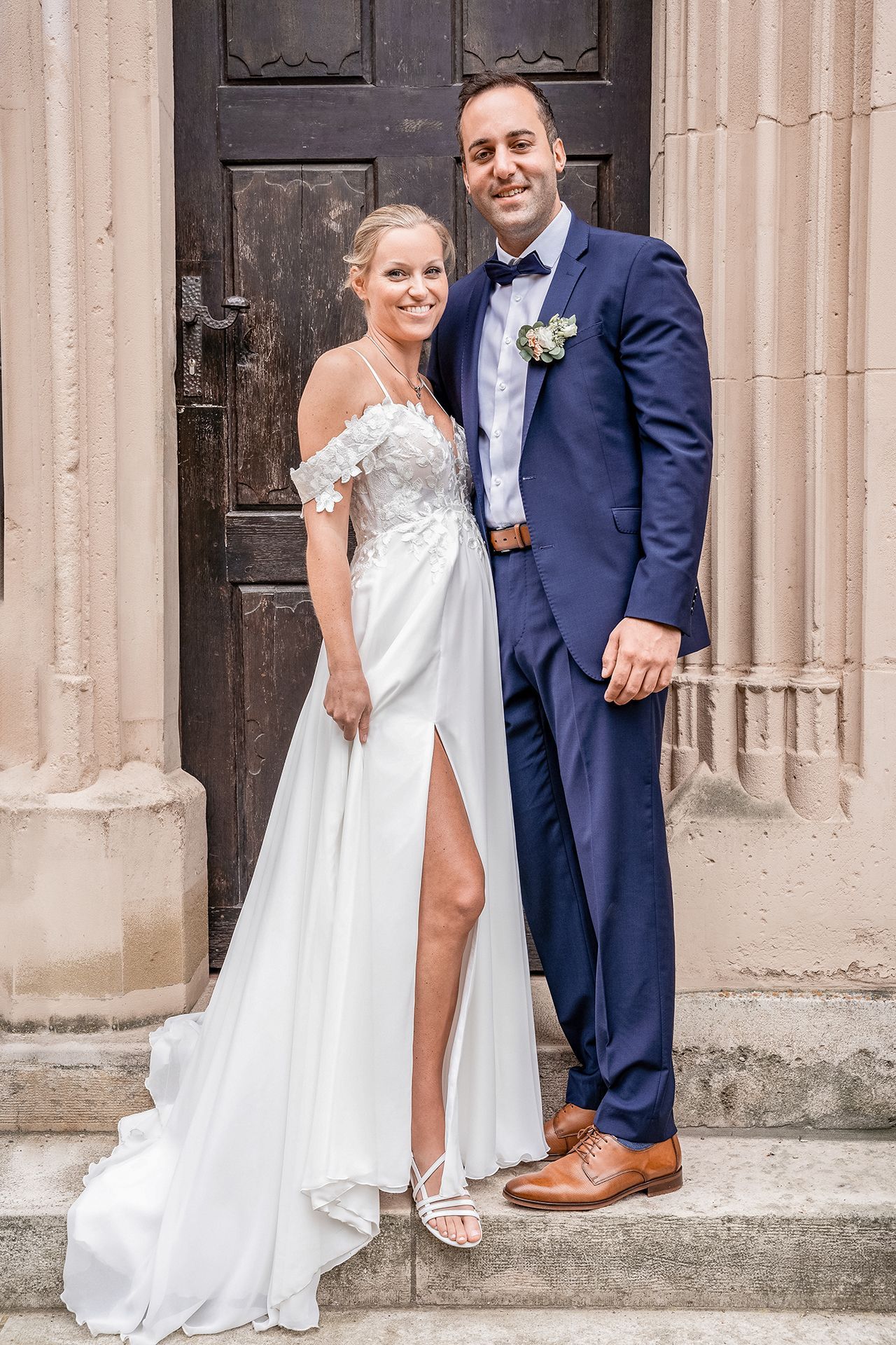 Brautpaar steht vor der Kirche und lächelt in die Kamera für Hochzeitsfotos