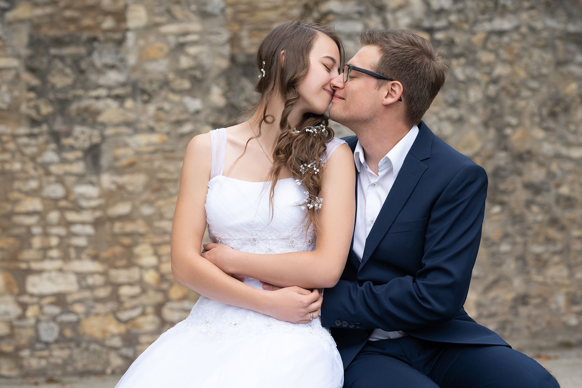 Braut sitzt auf Schoß des Bräutigams, sie küssen sich vor Steinmauer – Hochzeitsfotografie Mainz