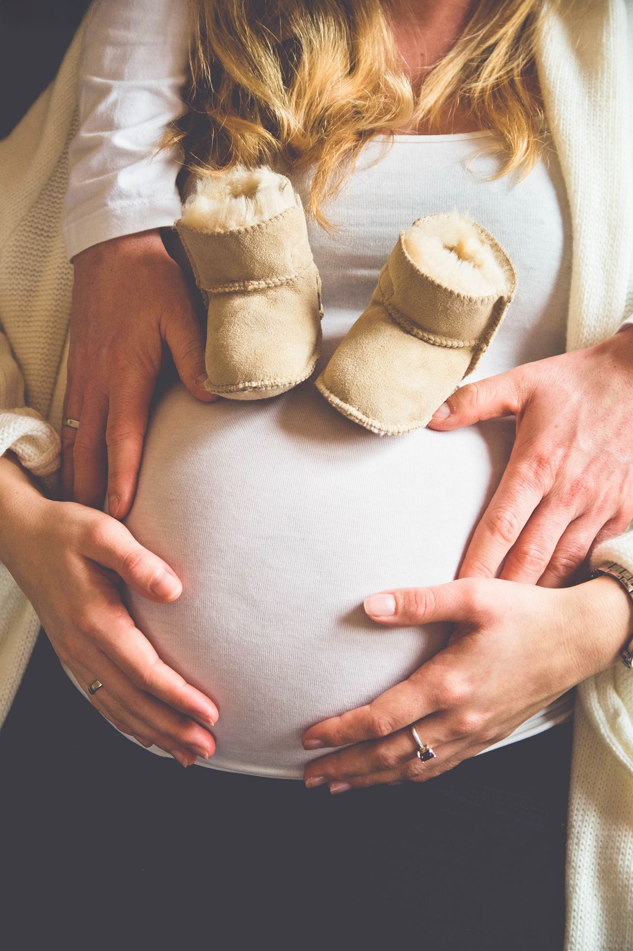 Babybauch im Fokus – Paar hält Hände am Bauch, Babyschuhe oberhalb des Bauches