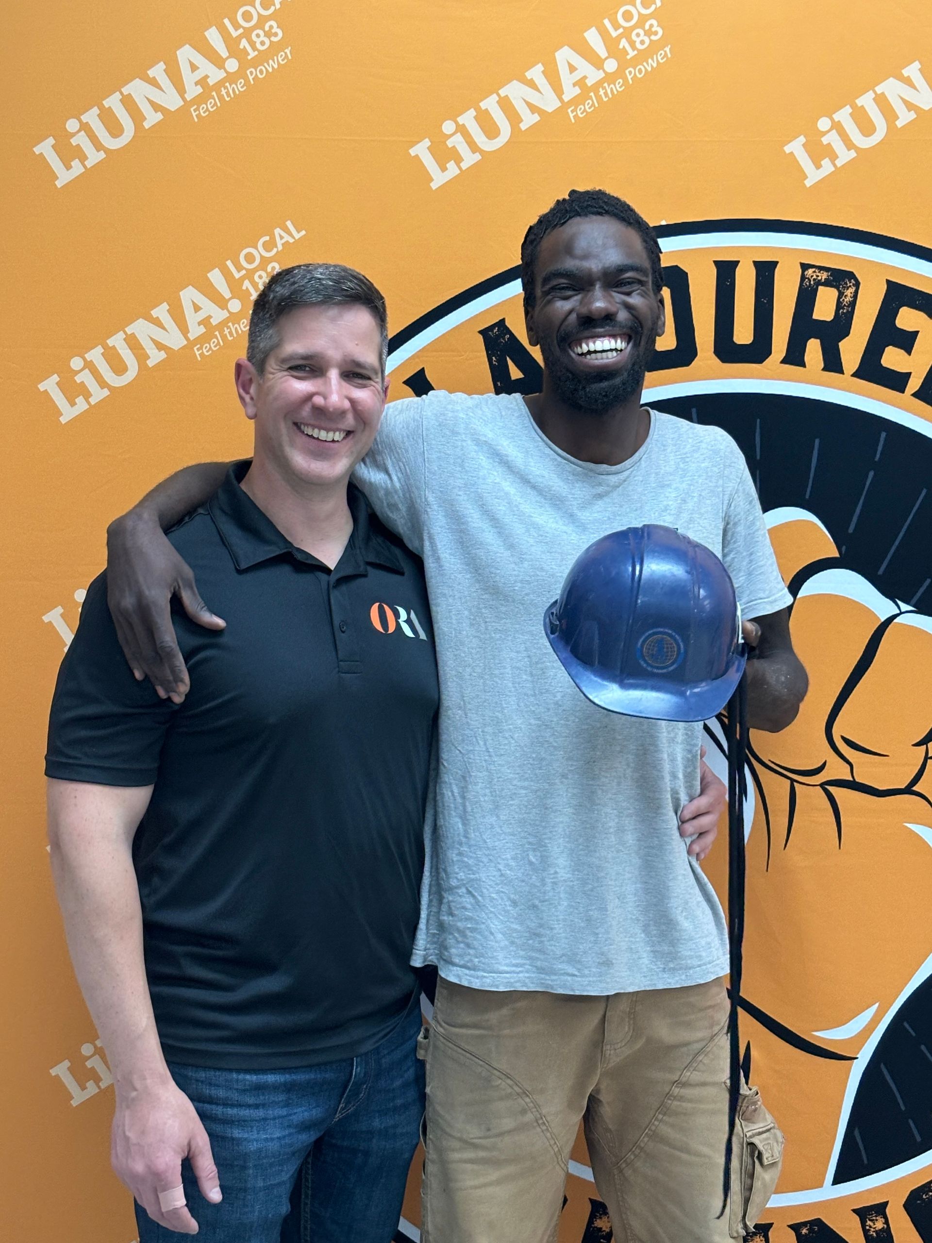 Two men are standing next to each other and one of them is holding a hard hat.