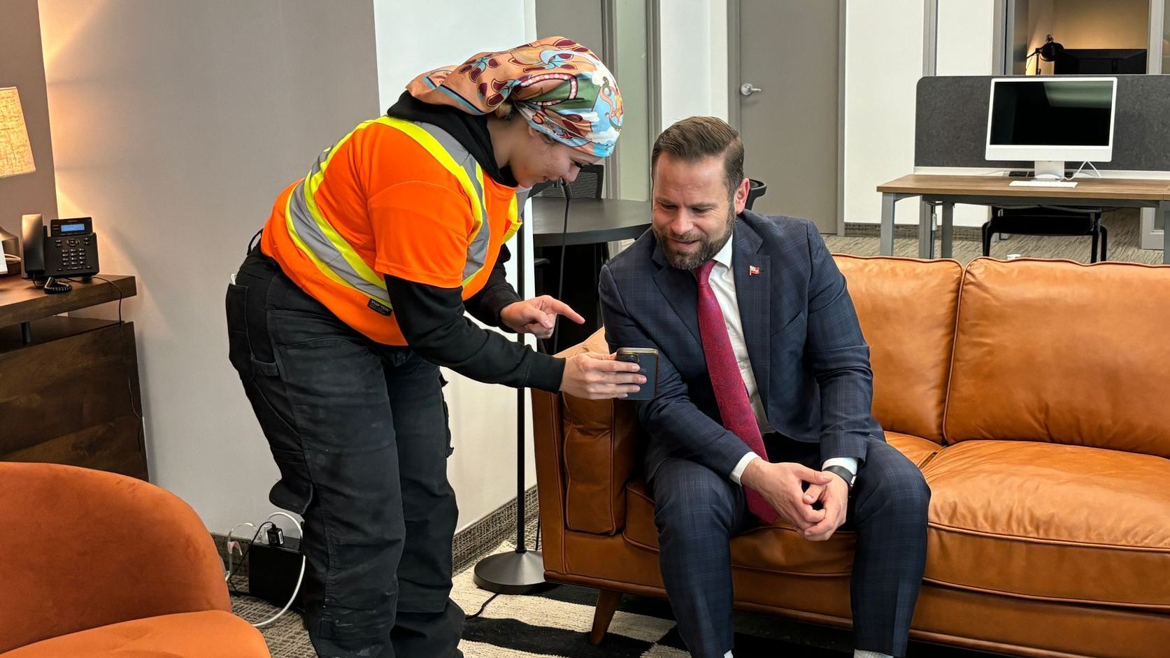 A female construction worker sharing pictures on her phone to a man in a suit and tie is sitting on a couch.