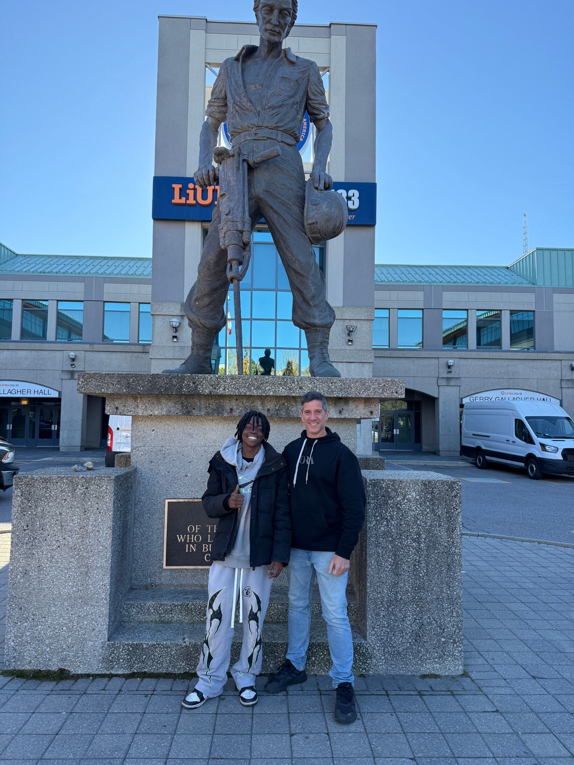 Two men are standing in front of a statue of a man holding a helmet.