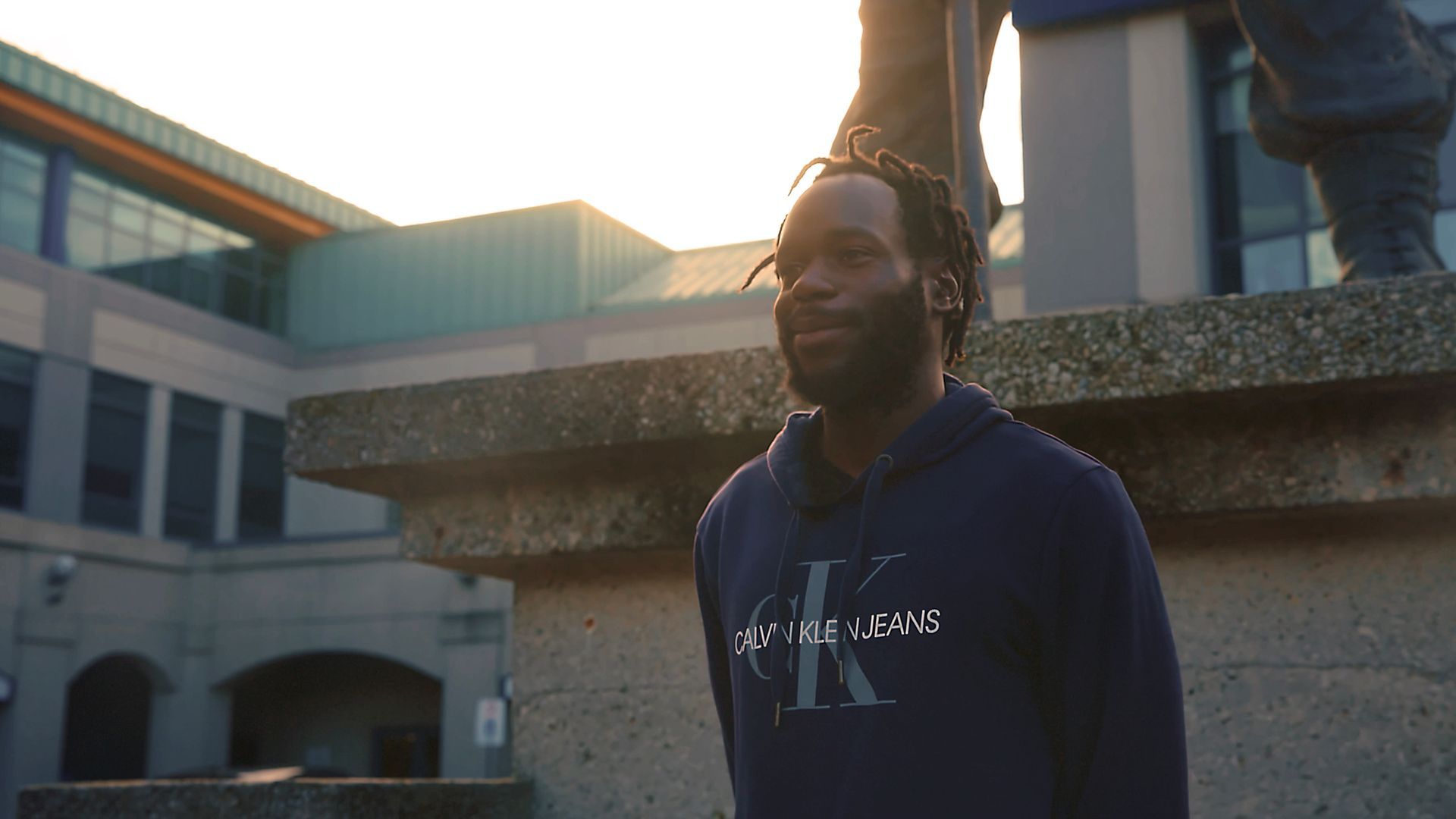 Man wearing a blue Calvin Klein hoodie, standing outside a building in sunlight.
