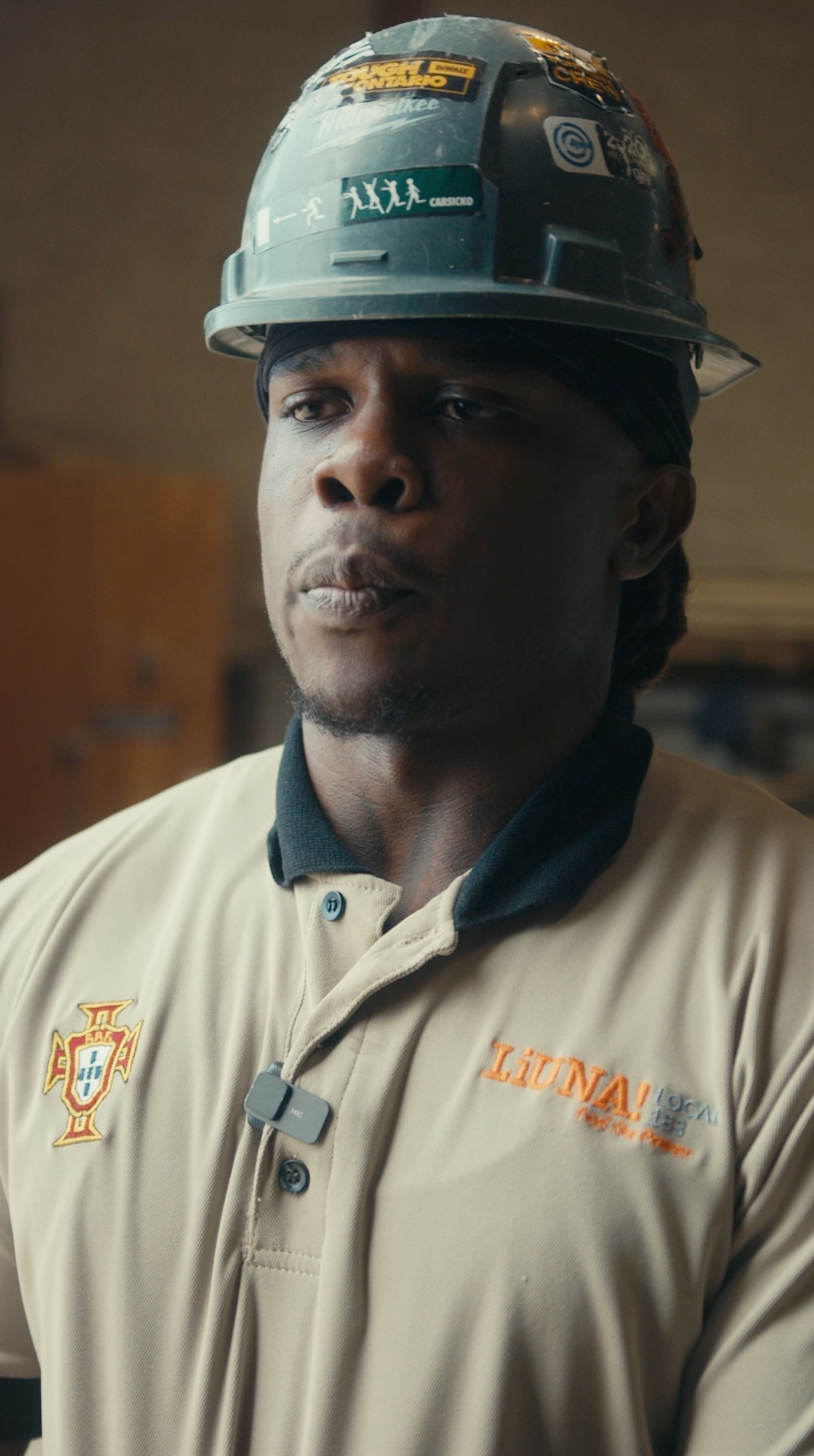 A Black man wearing a work uniform and a hard hat looks slightly off-camera in a factory setting.