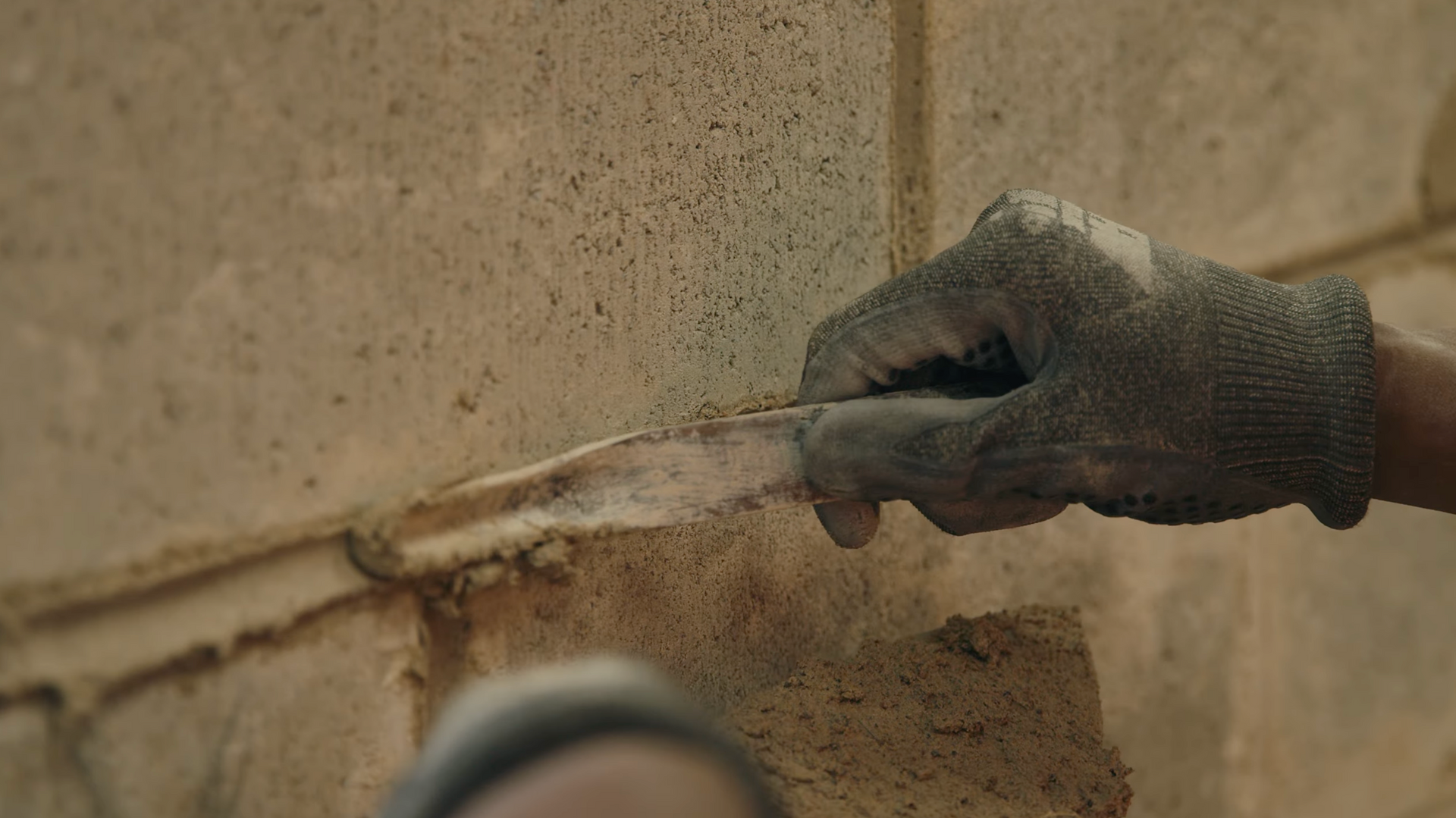 Hand wearing a glove smoothing mortar between a brick wall joint.