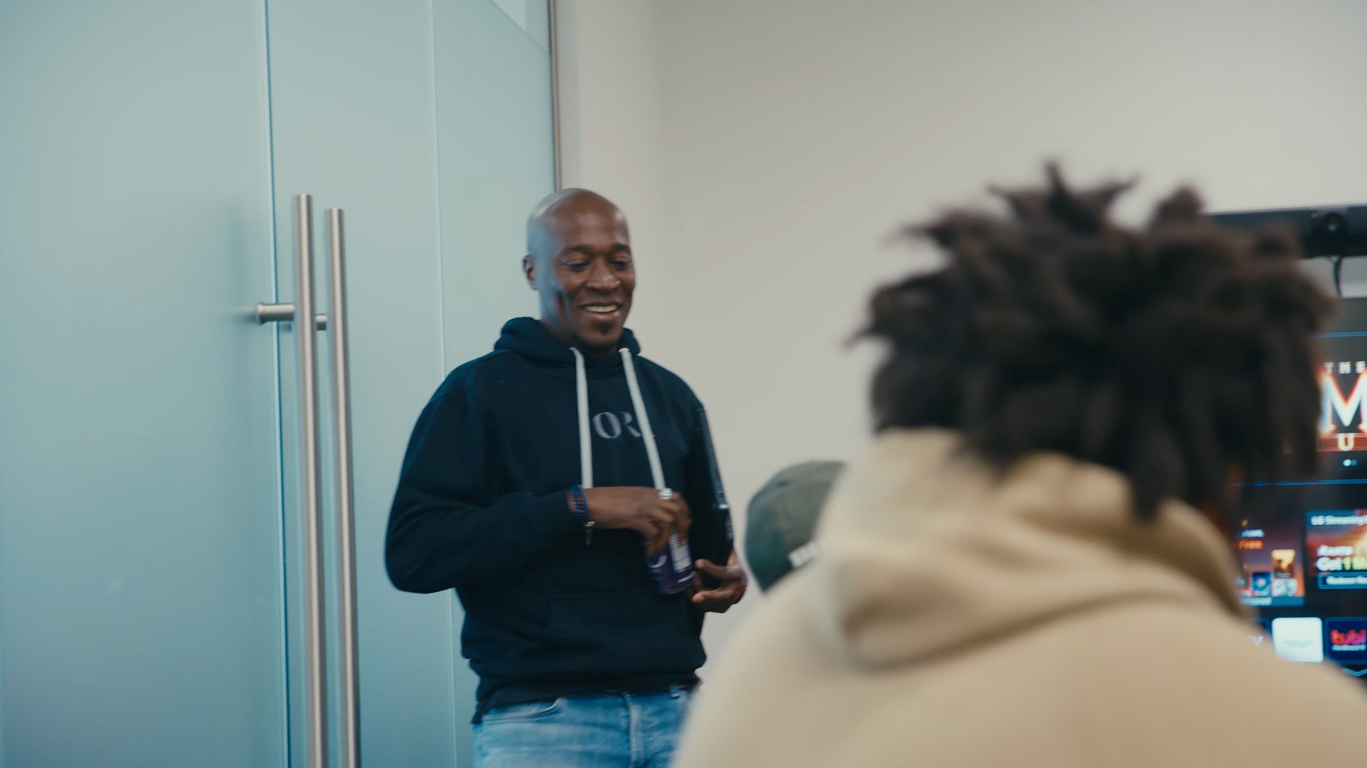 Man in hoodie smiles, gestures while speaking in a room with a clear glass wall and person facing away.