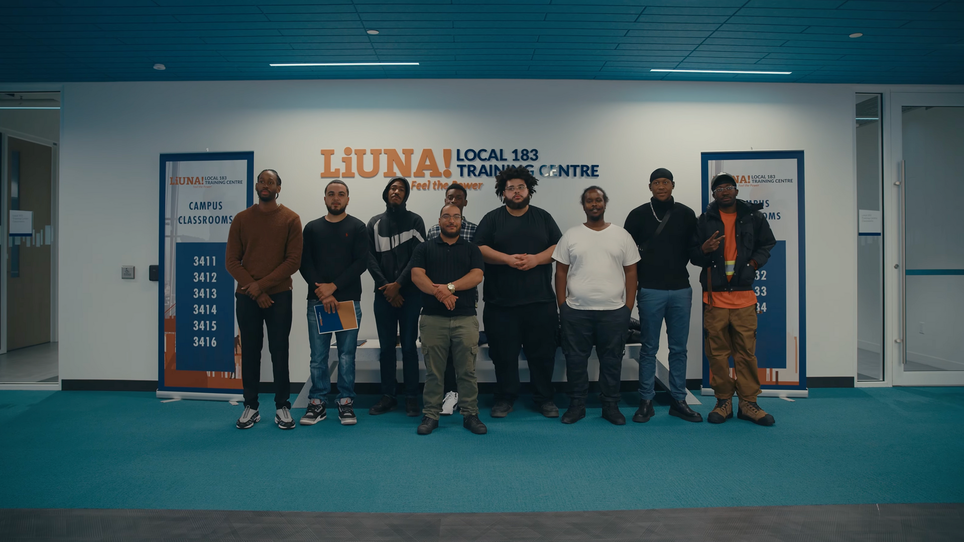 Group of ten people stand in front of a building with a blue wall. Signs with writing are above the group.